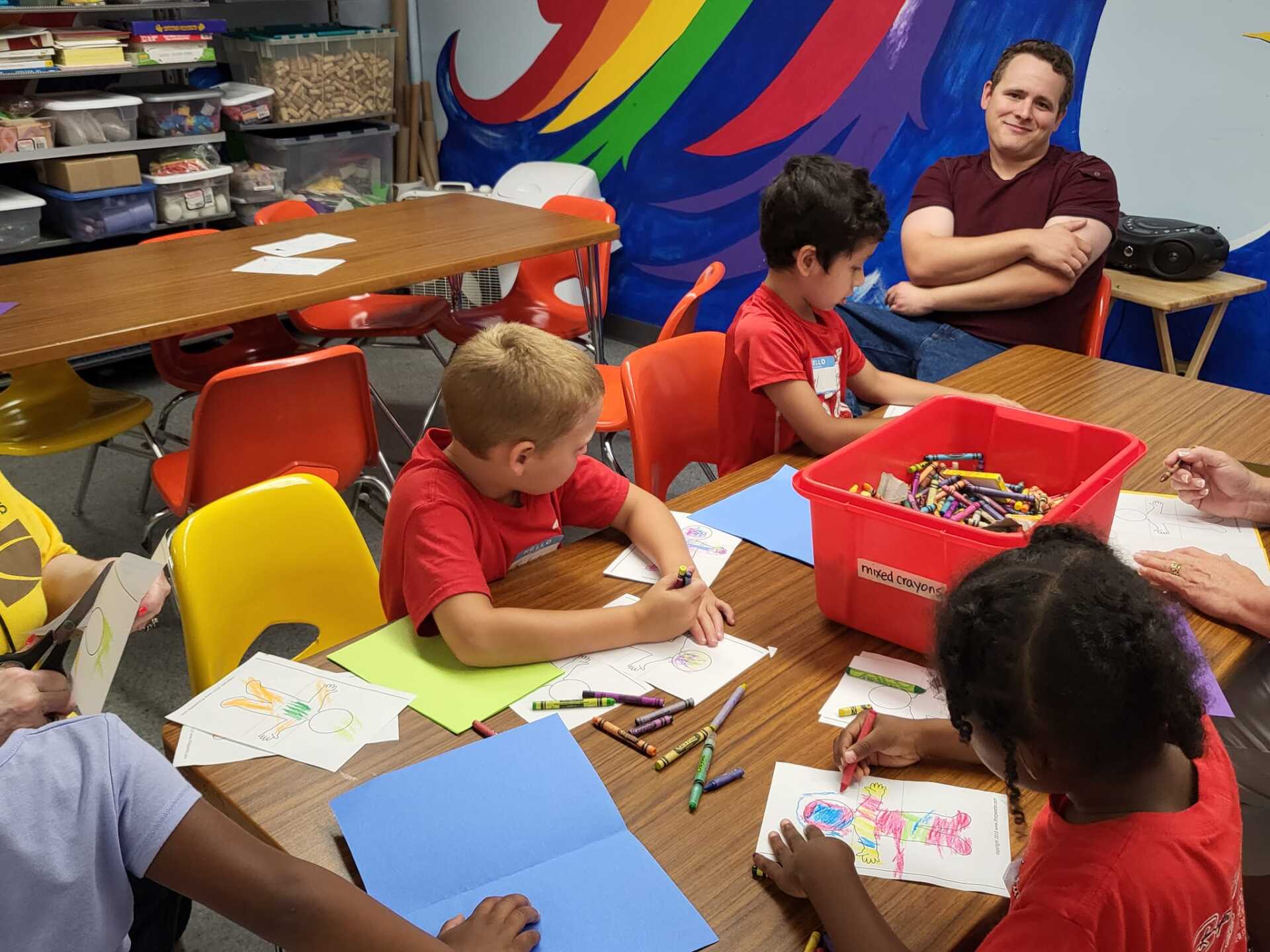 Children drawing at tables in a colorful classroom, with an adult supervising nearby.