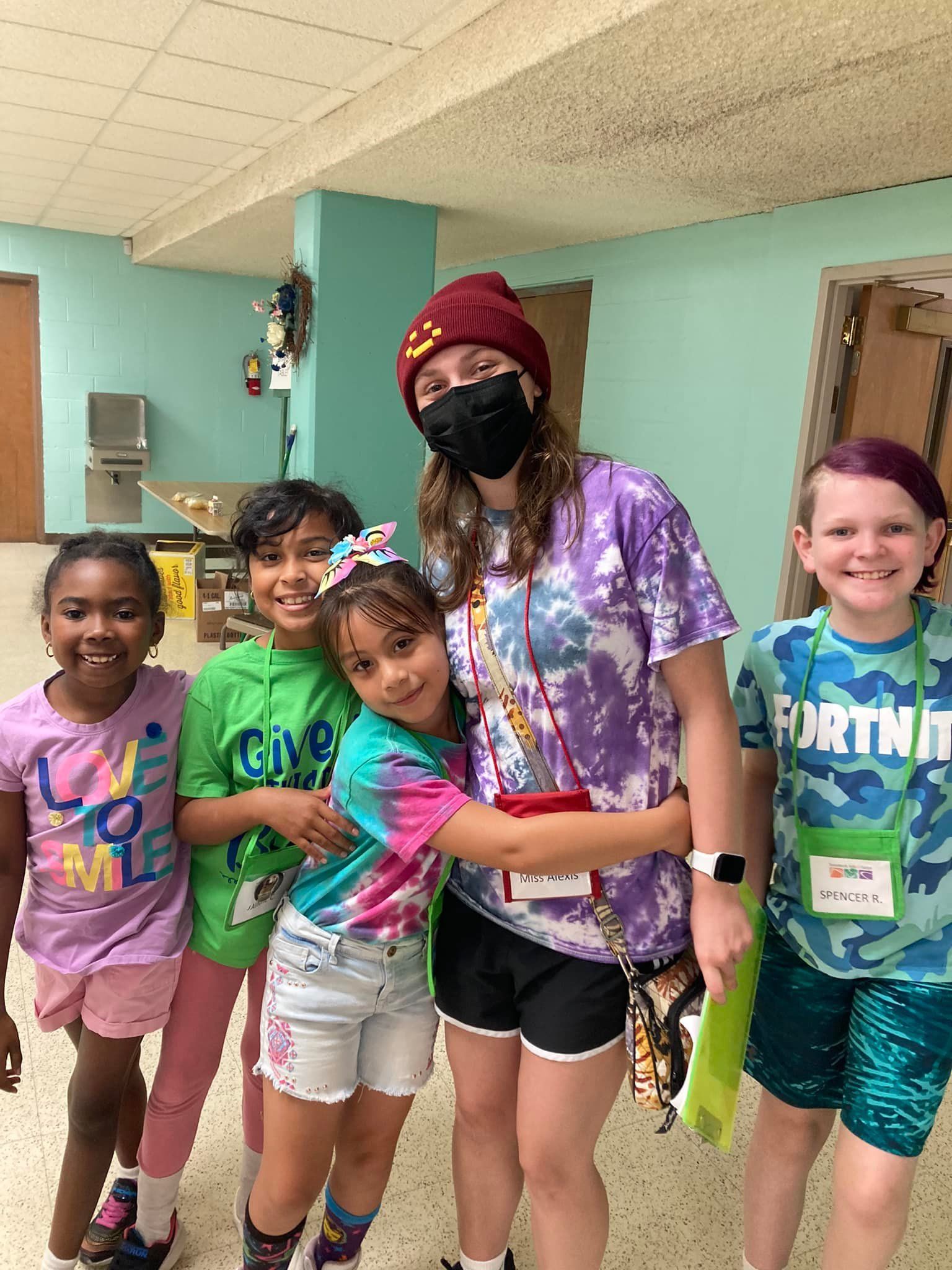 Five smiling children posing in a hallway, wearing colorful clothes and lanyards.