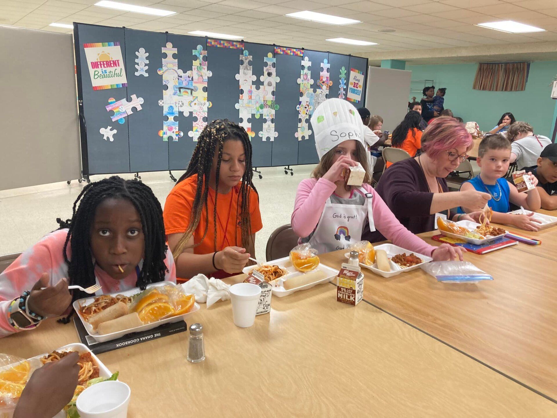 Children eating lunch together at a long table in a cafeteria, with trays, drinks, and drawings on the wall.