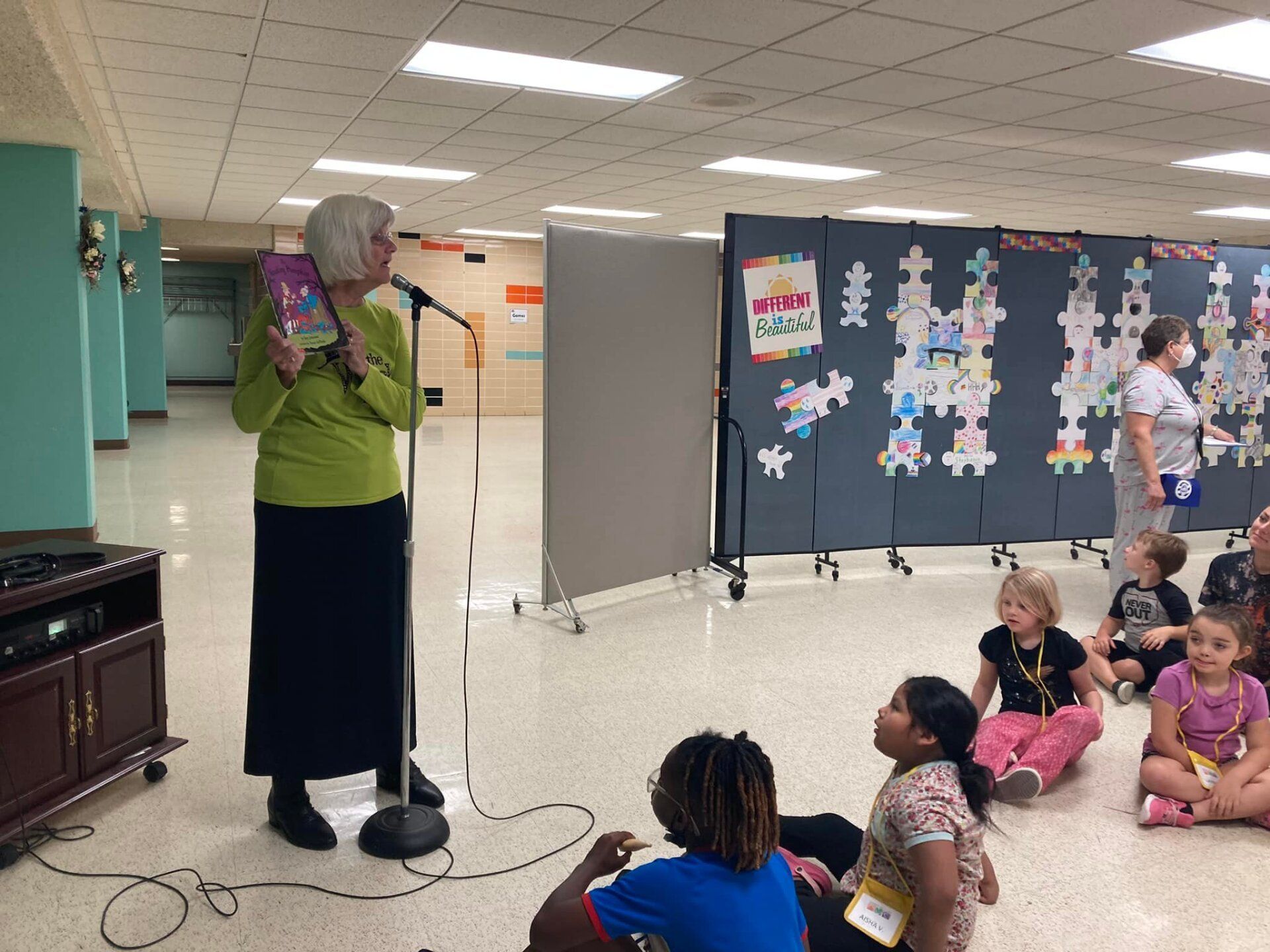Woman speaking into a microphone to seated children in a classroom or hallway with posters on the wall