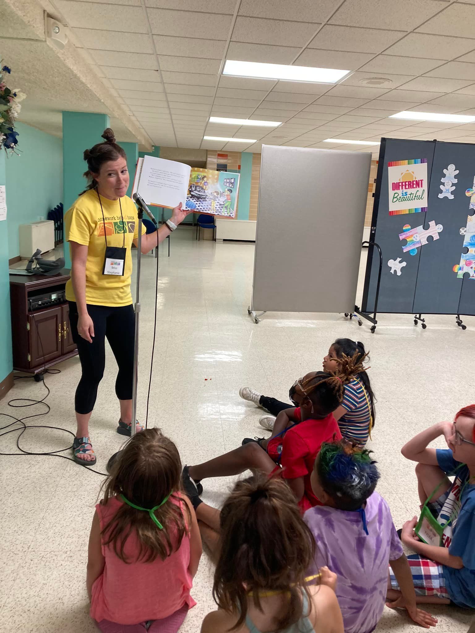 Teacher in yellow shirt speaks to seated children in a classroom hallway with display boards.