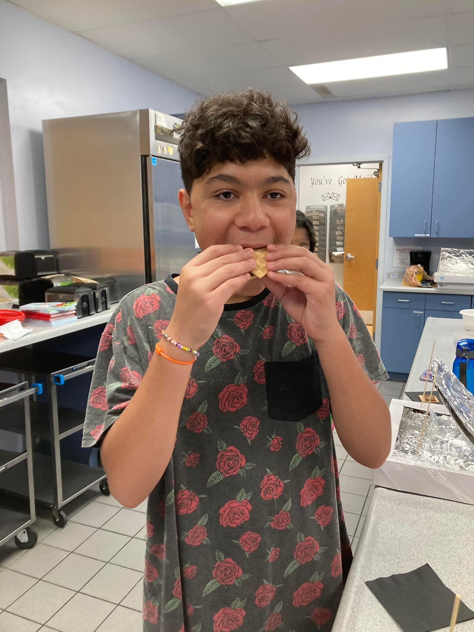 Person eating a sandwich in a kitchen, wearing a dark floral shirt.