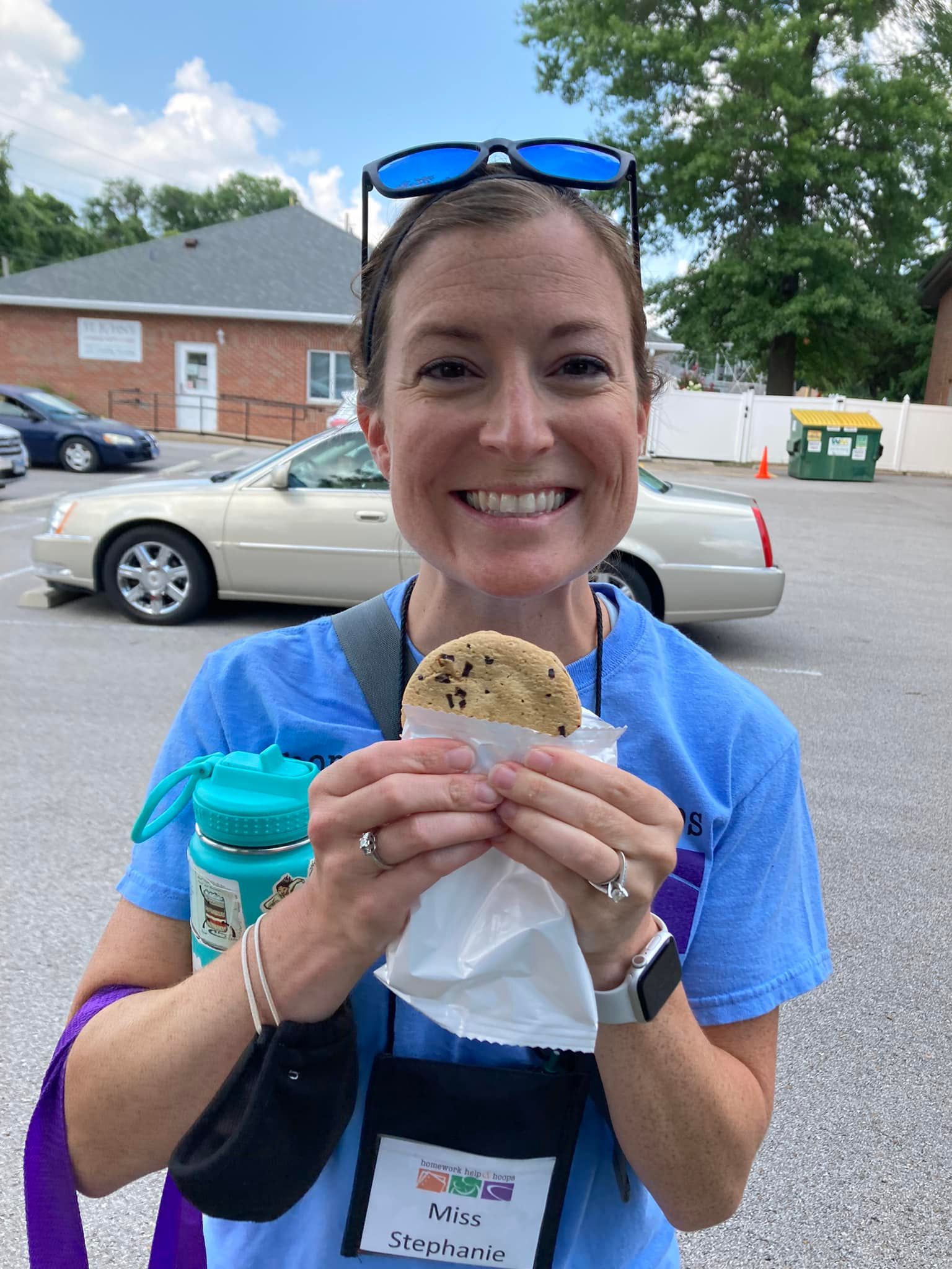 Woman in blue shirt holding a cookie and drink, smiling outdoors in a parking lot.