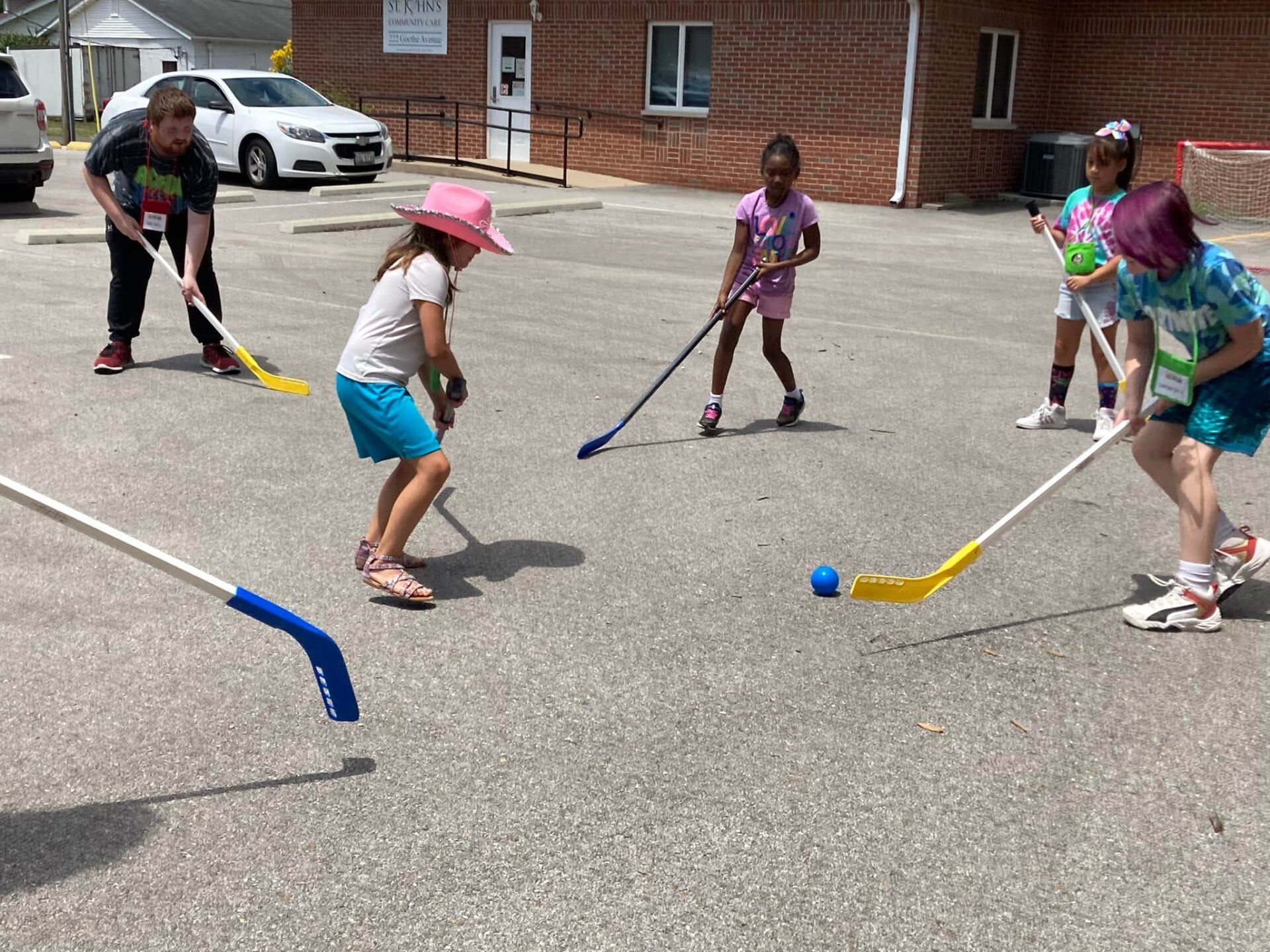 Children playing street hockey with colorful sticks and a blue ball outside a brick building