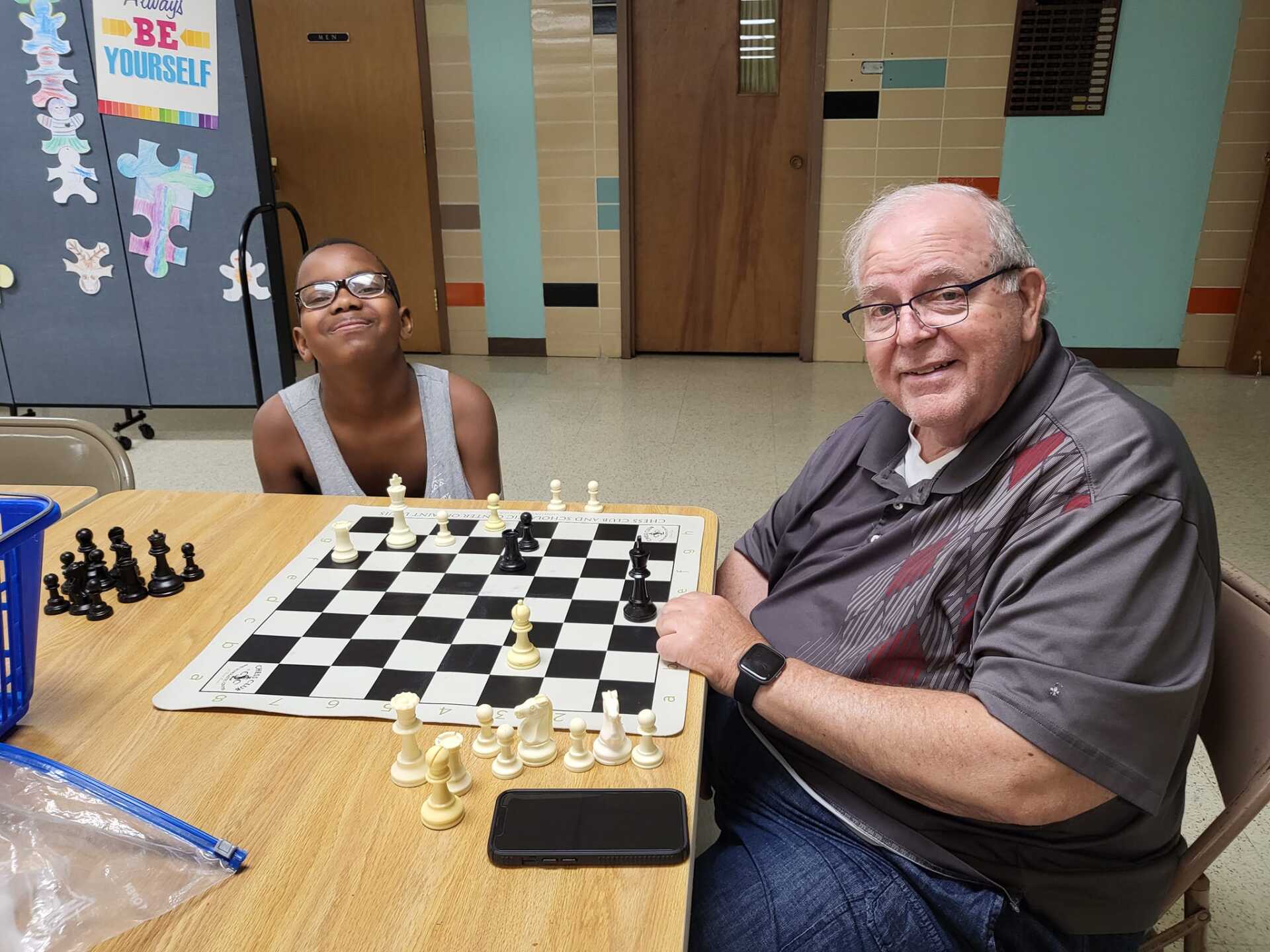 Two people seated at a table, smiling beside a chessboard in a room.