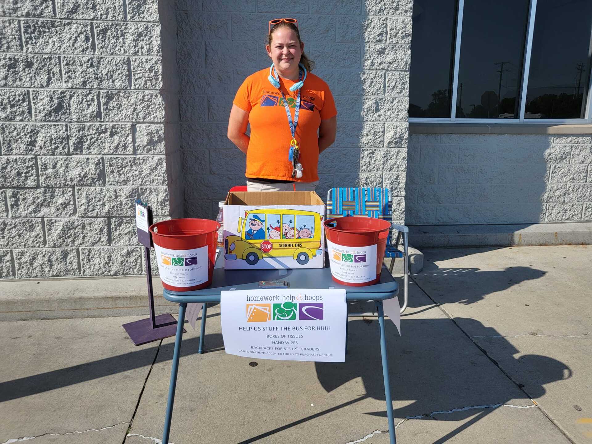 Person in orange shirt standing behind a table with donation buckets and signs outside a building