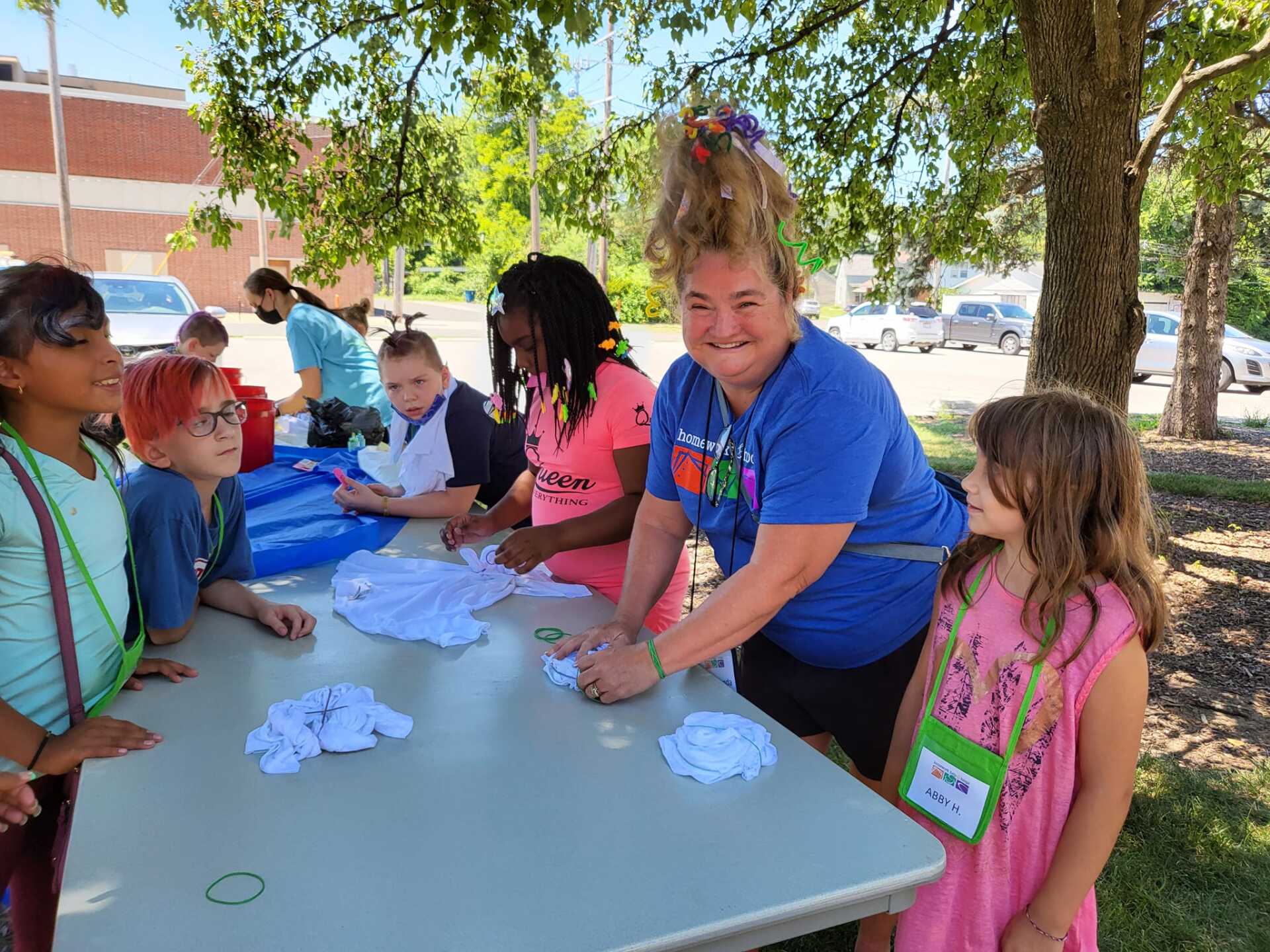 Children and adults making crafts at a picnic table outdoors under trees