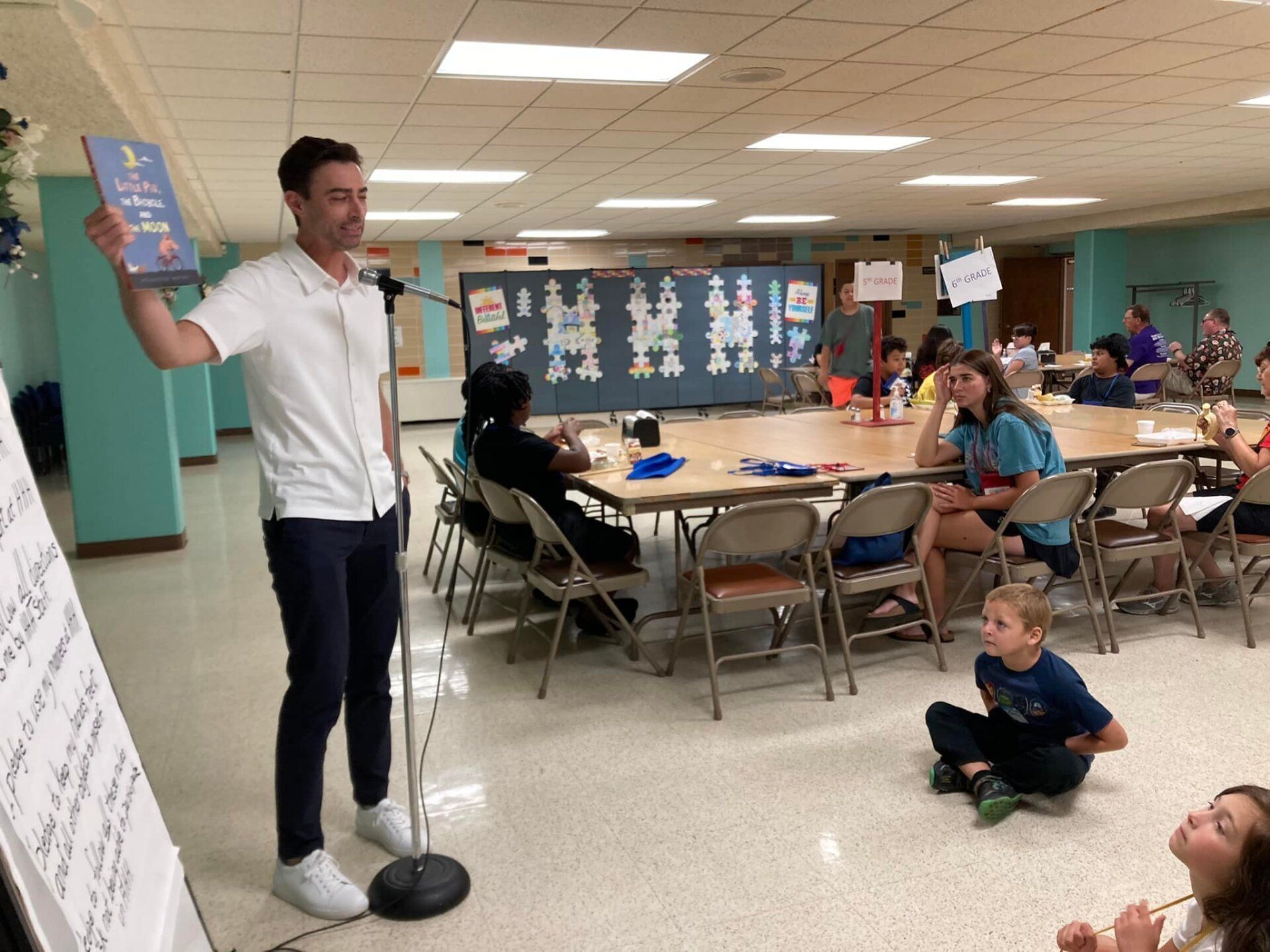 Man speaking into a microphone in a cafeteria while seated children listen at tables and on the floor.