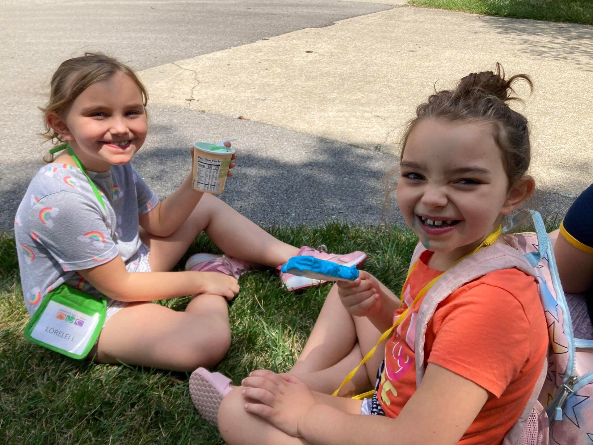 Two children sitting on grass, smiling and holding snacks in a sunny outdoor area.