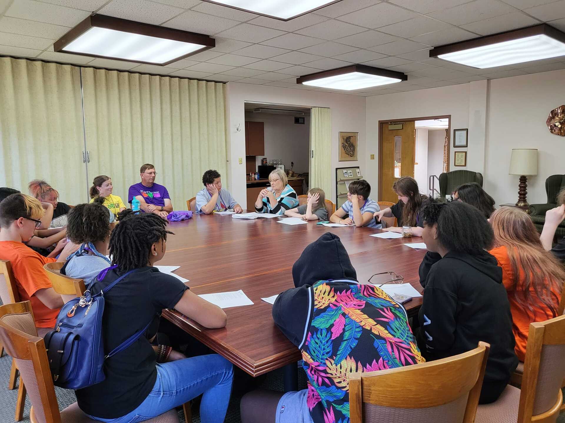 People seated around a conference table in a meeting room, discussing together under fluorescent lights.