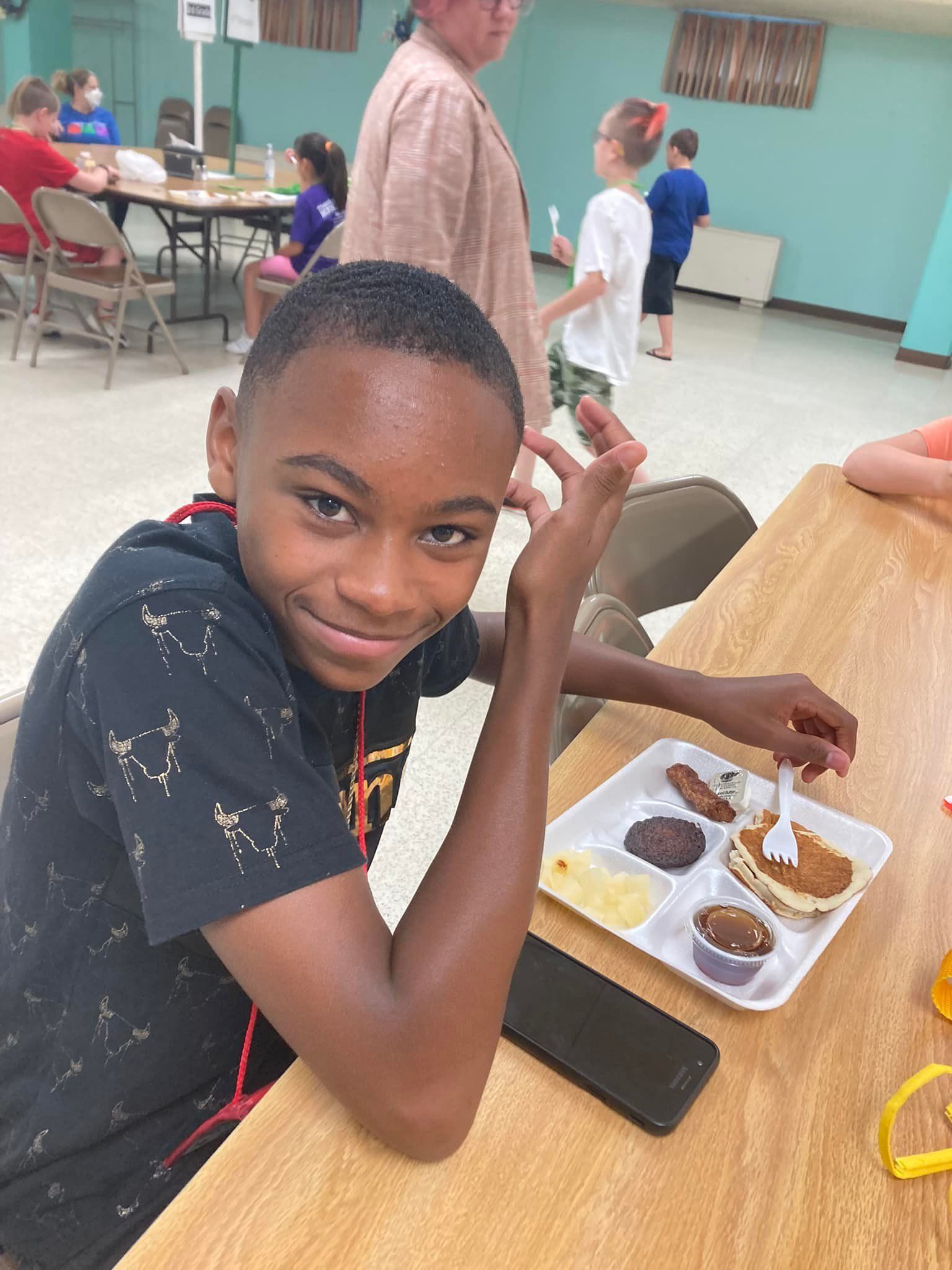 Child smiling at a cafeteria table, eating from a tray with assorted food and a phone beside it.