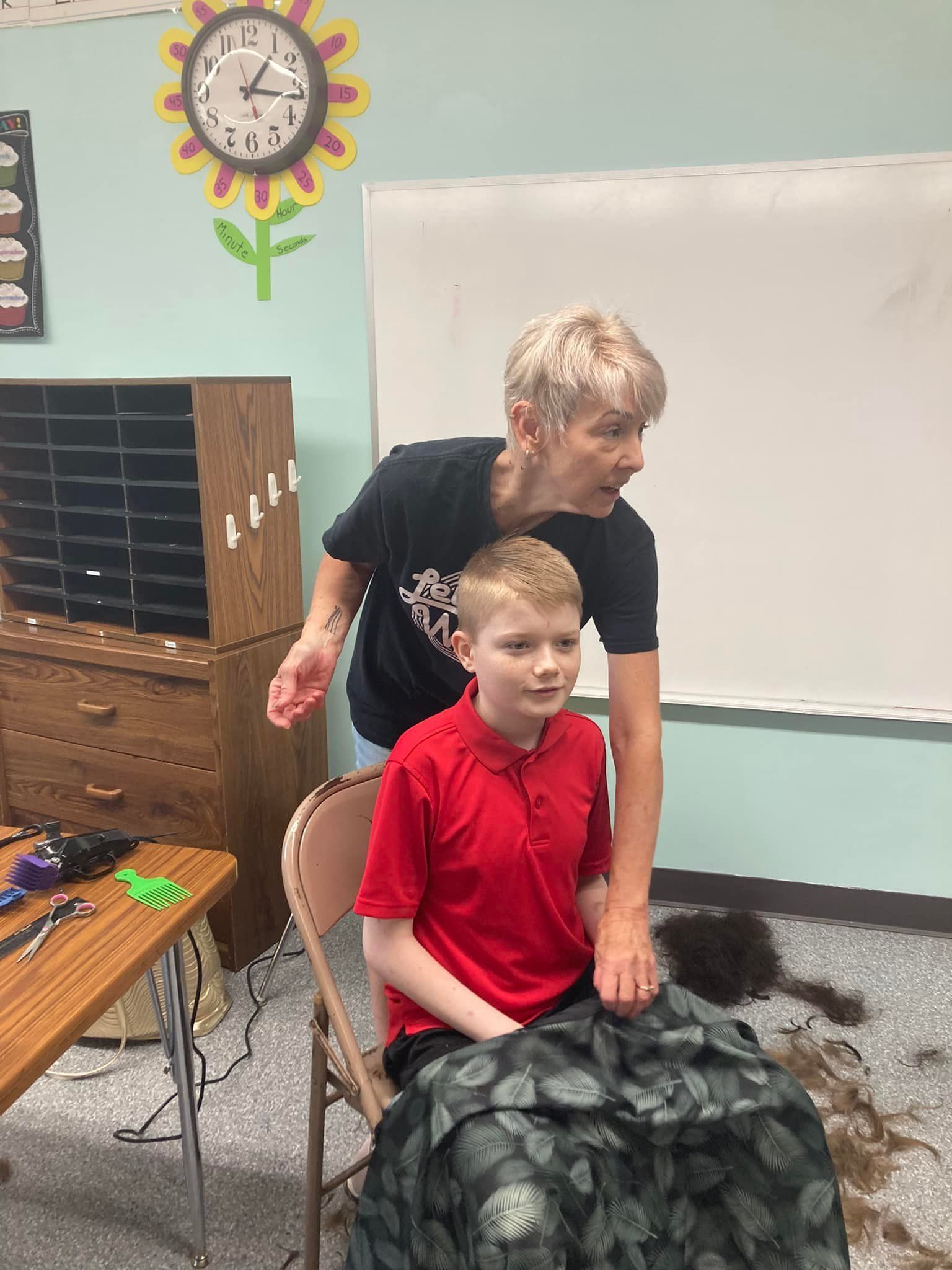 Teacher stands beside seated child in a classroom, near a whiteboard, clock, and table.