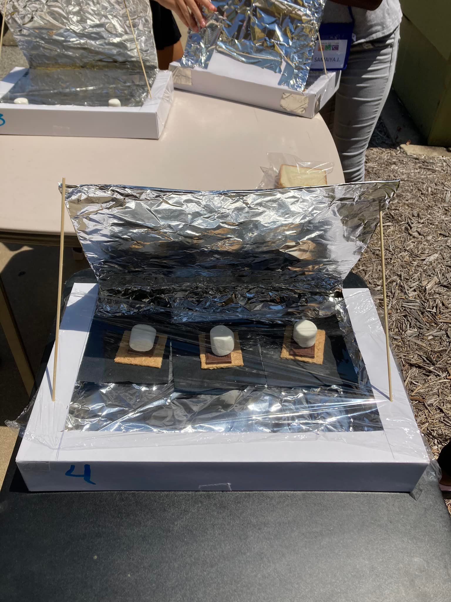 Solar cooker made from a cardboard box and foil, with three jars inside on gravel outdoors.