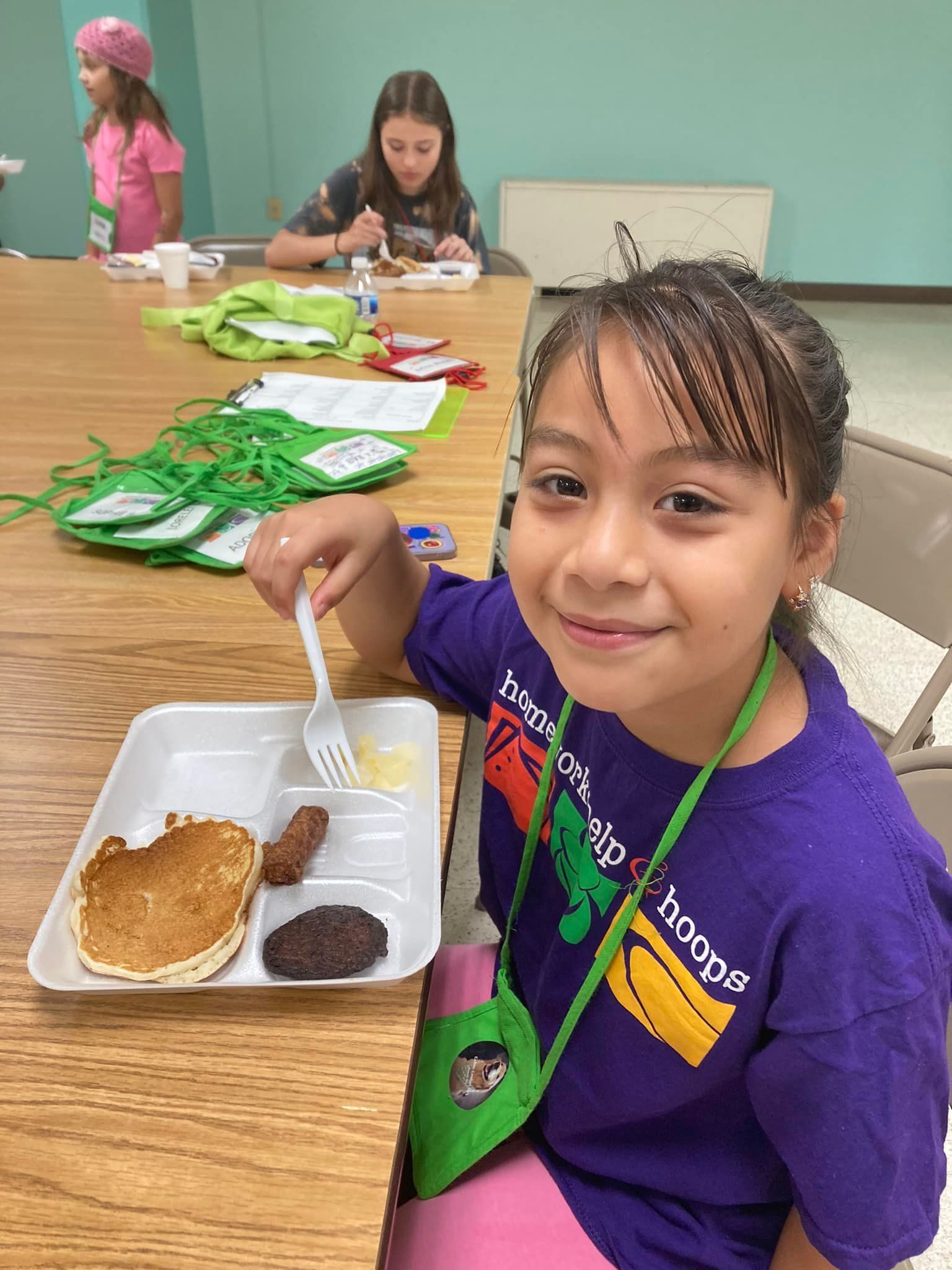 Child holding a cafeteria tray with a pancake and syrup at a table in a classroom lunch setting