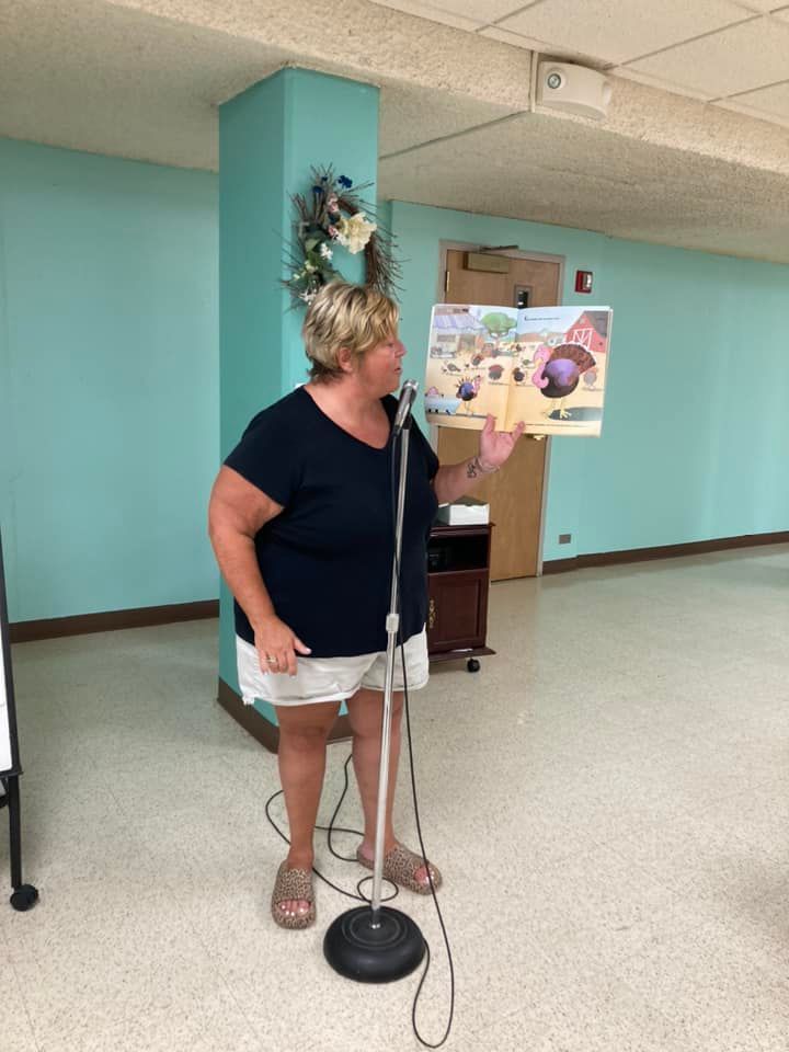 Woman speaking into a microphone in a teal community room, standing beside a poster board