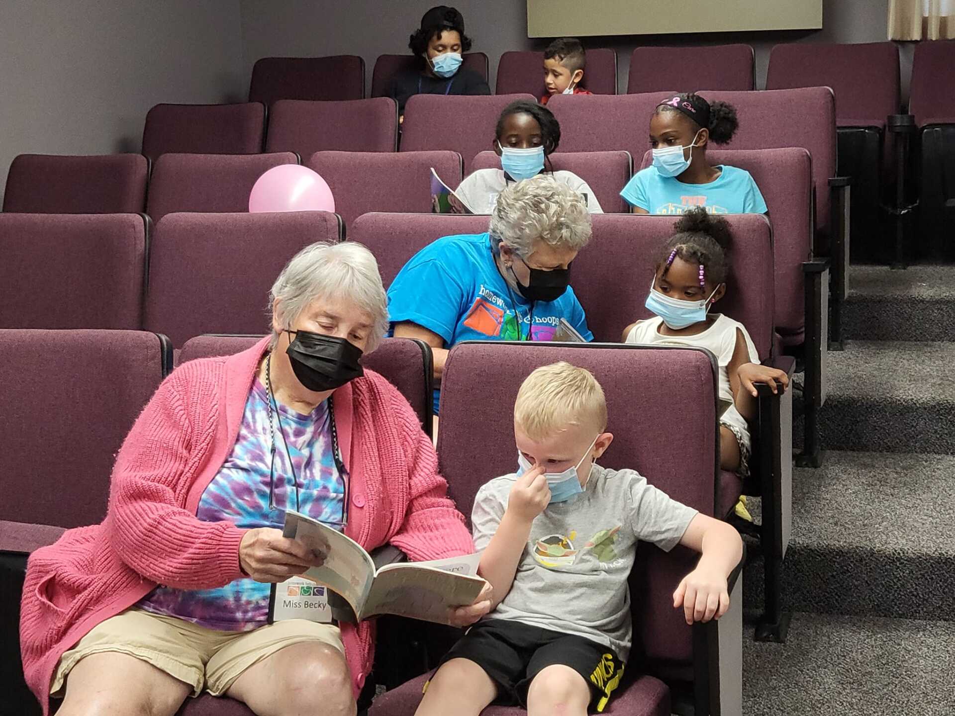 People seated in theater chairs, some wearing masks and reading together.