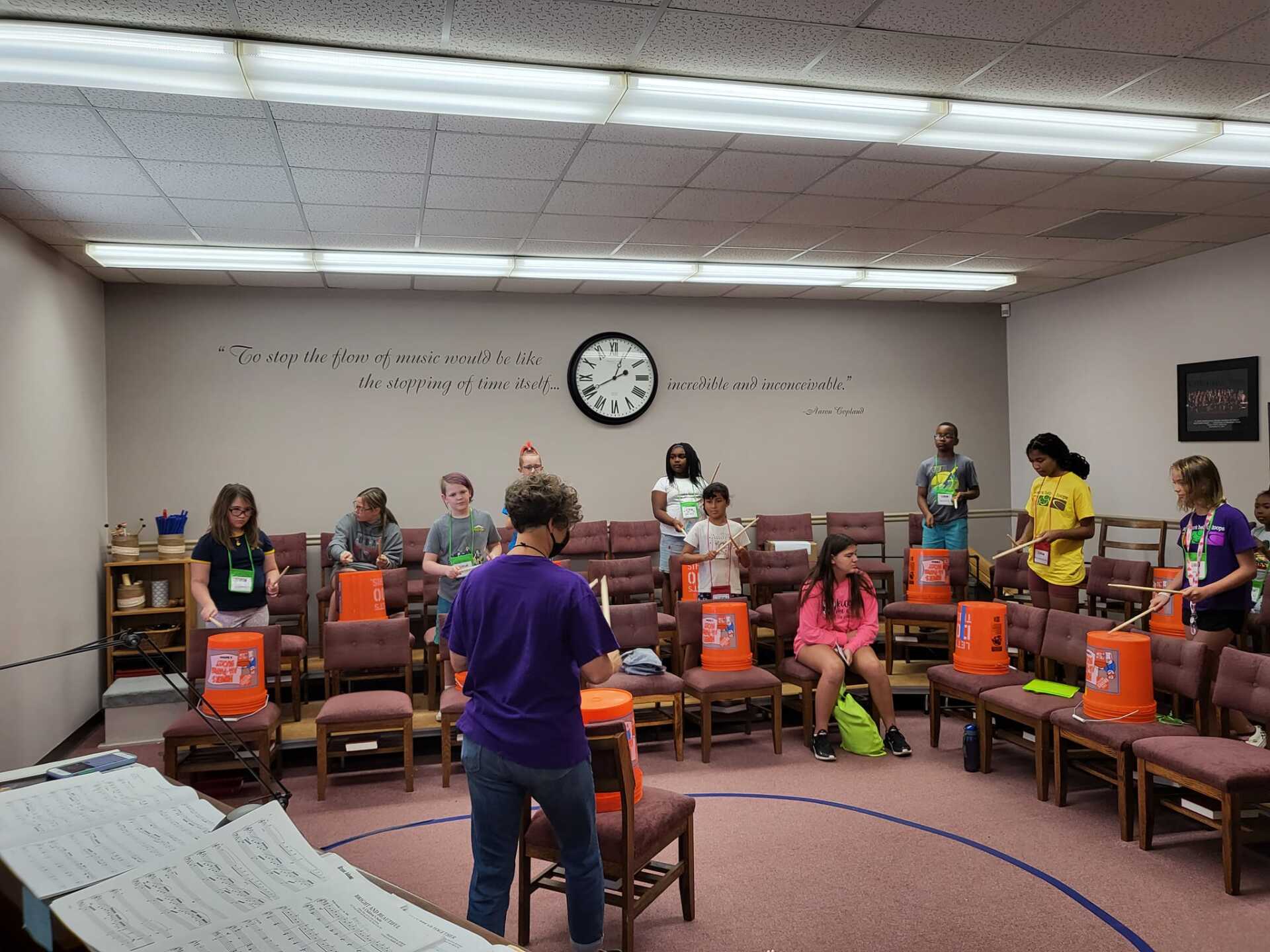 Group standing and sitting in a classroom or meeting room, facing a speaker near the front wall clock.