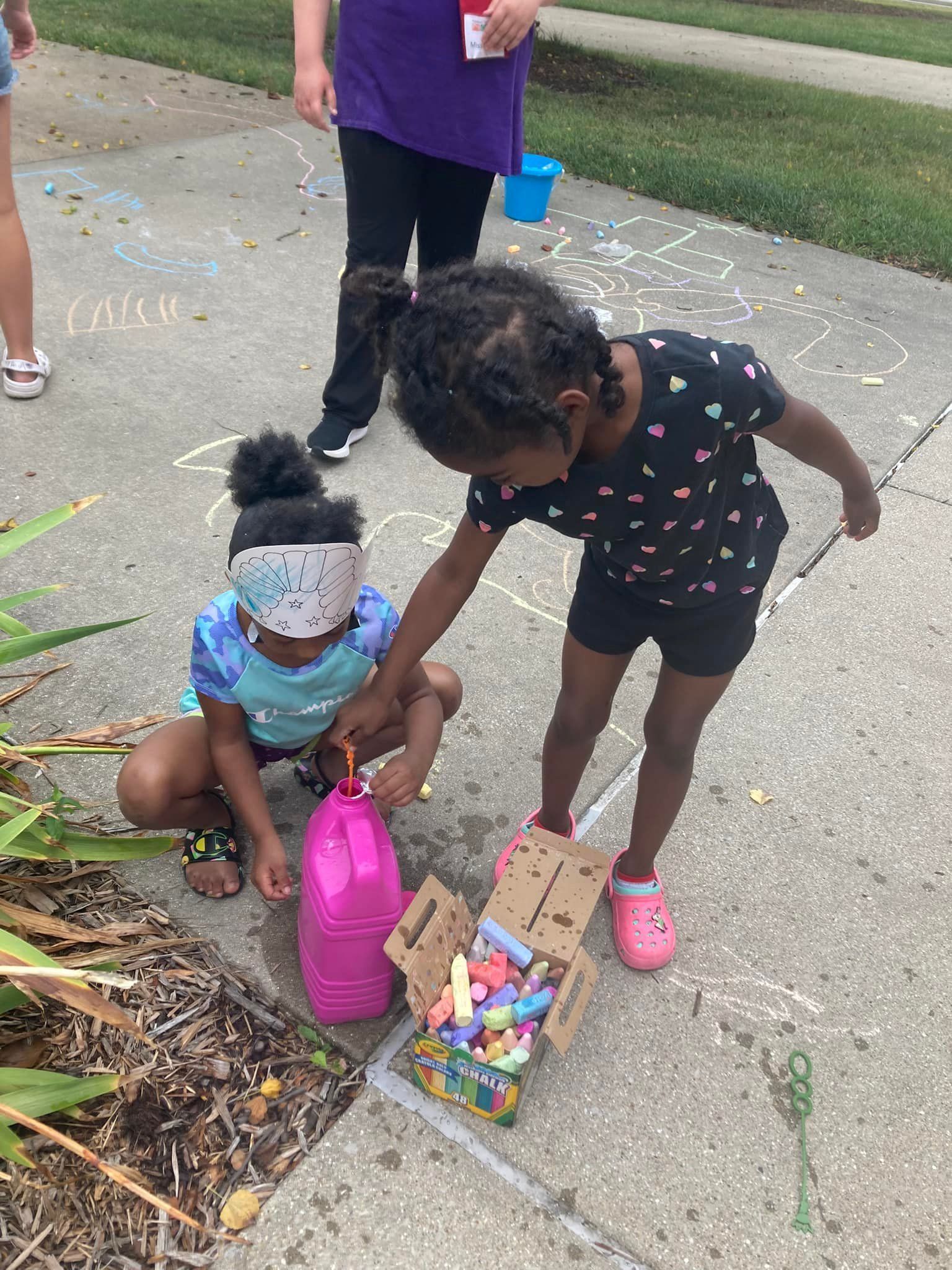 Two children on a sidewalk reach for a pink bag and a box of small toys next to them.