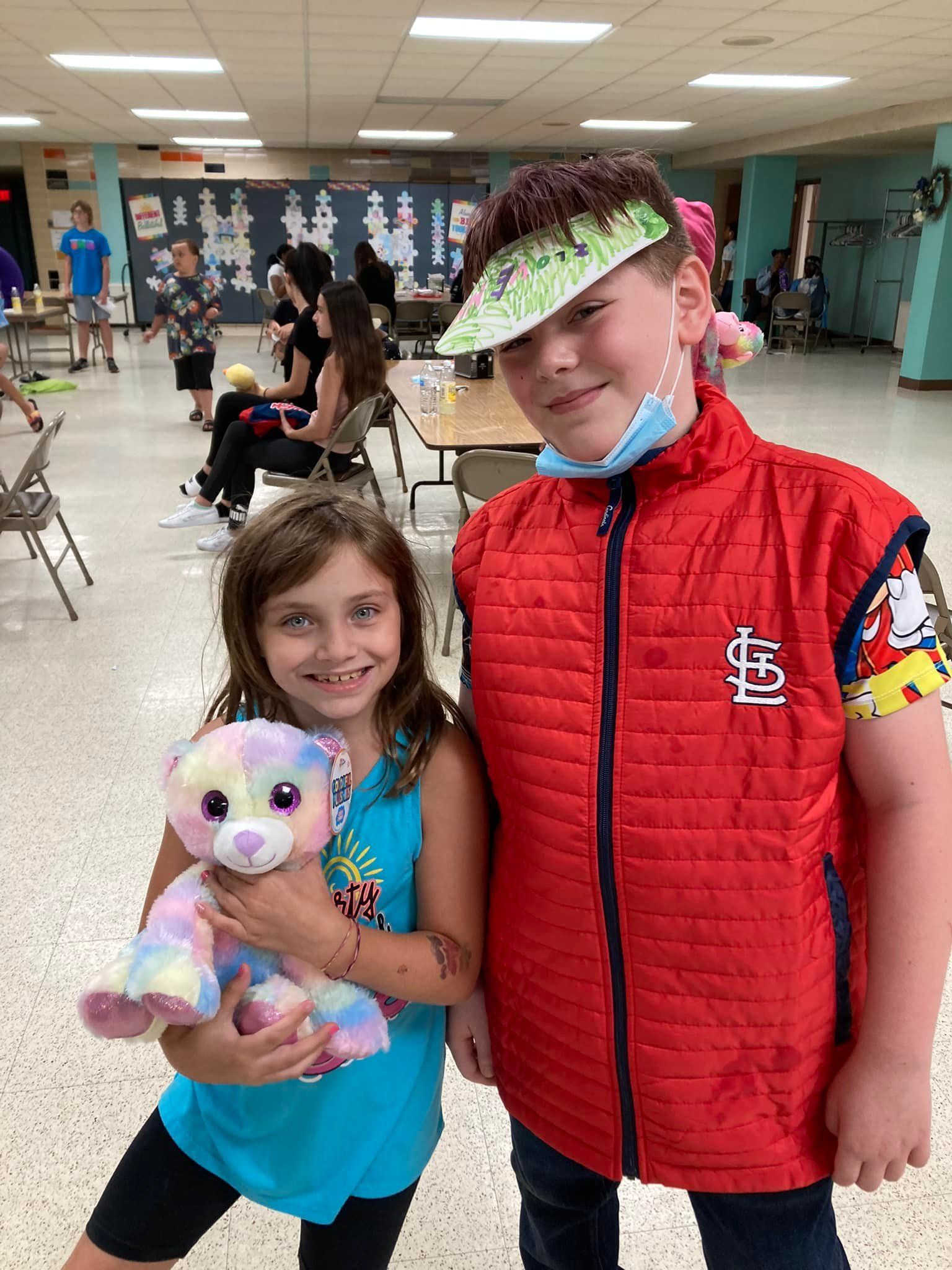 Two children smiling indoors; one holds a plush toy and the other wears a red vest and visor.