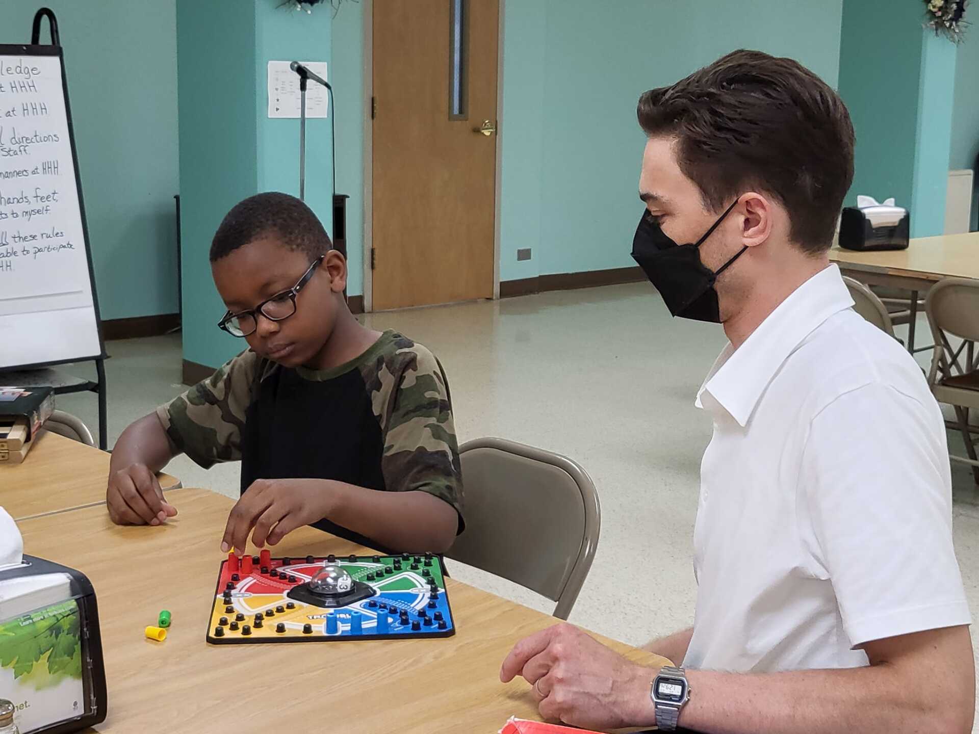 Two people sit at a table, one working with colorful materials while the other watches in a classroom.