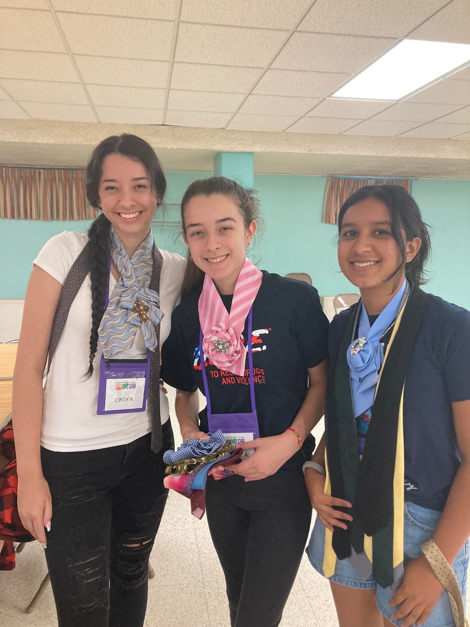 Three people smiling indoors, standing together and holding medals or ribbons in a school hallway