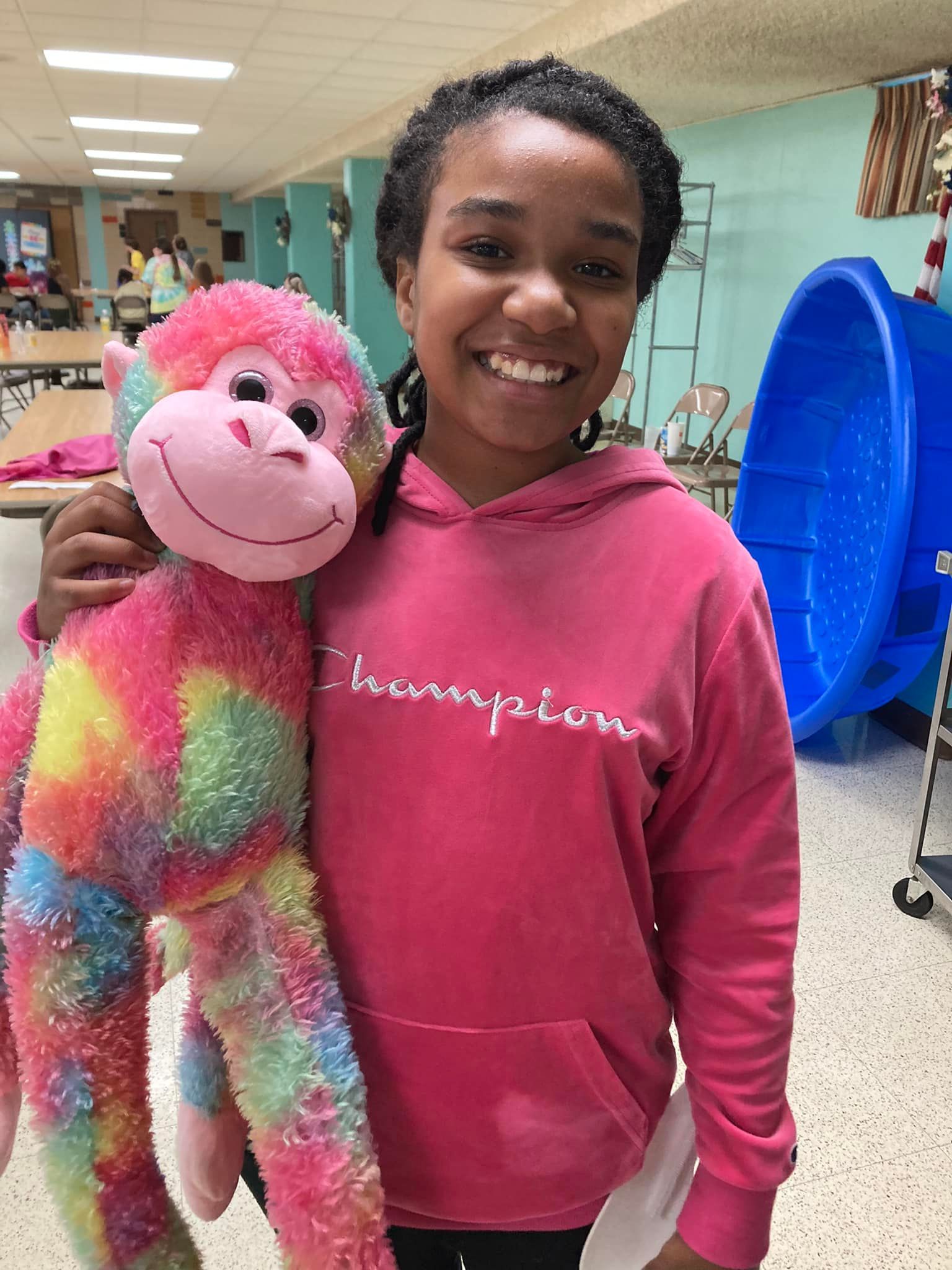 Smiling student holding a bright pink and rainbow stuffed monkey in a school hallway