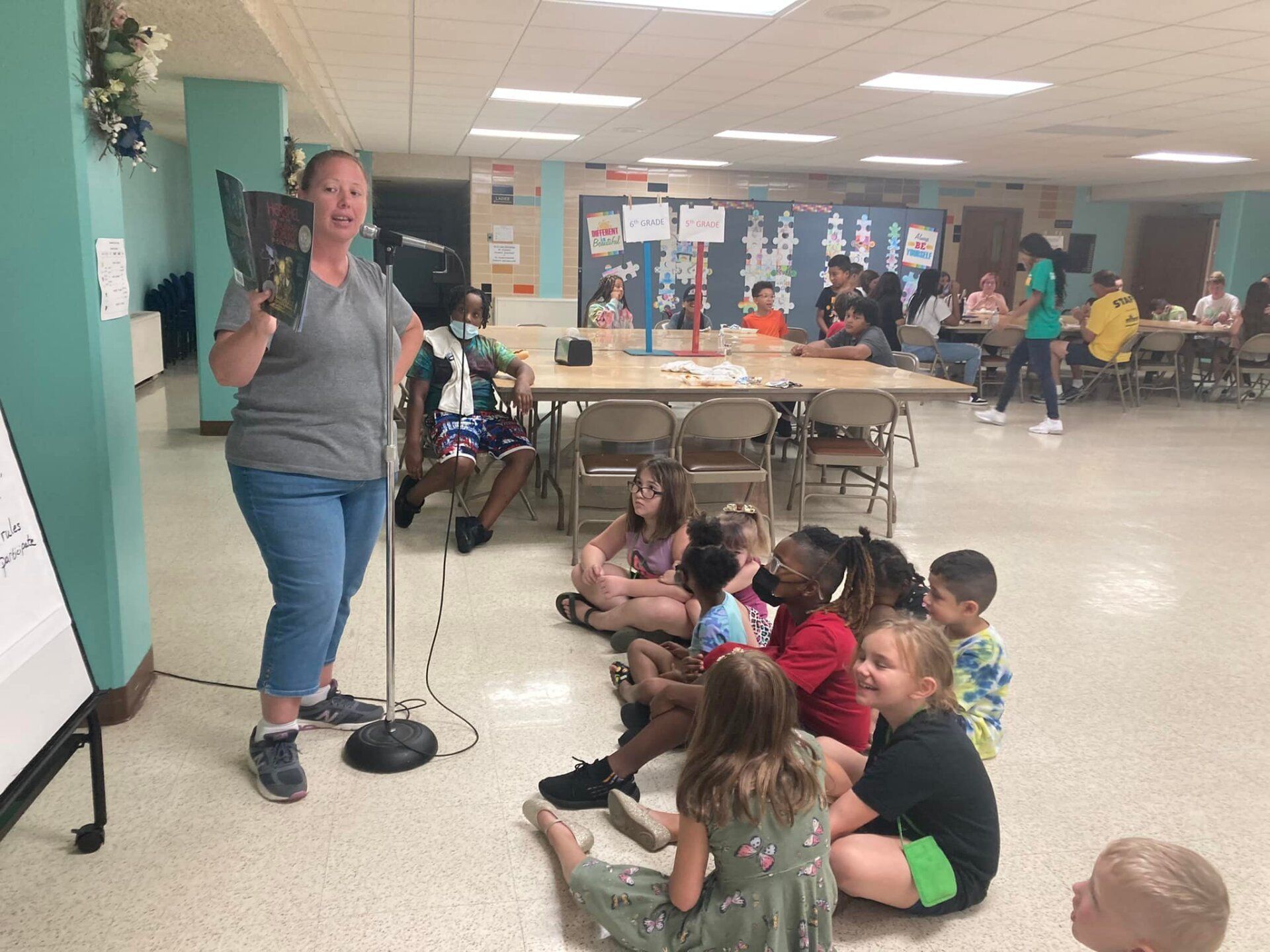 Teacher speaking to seated children in a school cafeteria with a projector screen nearby.