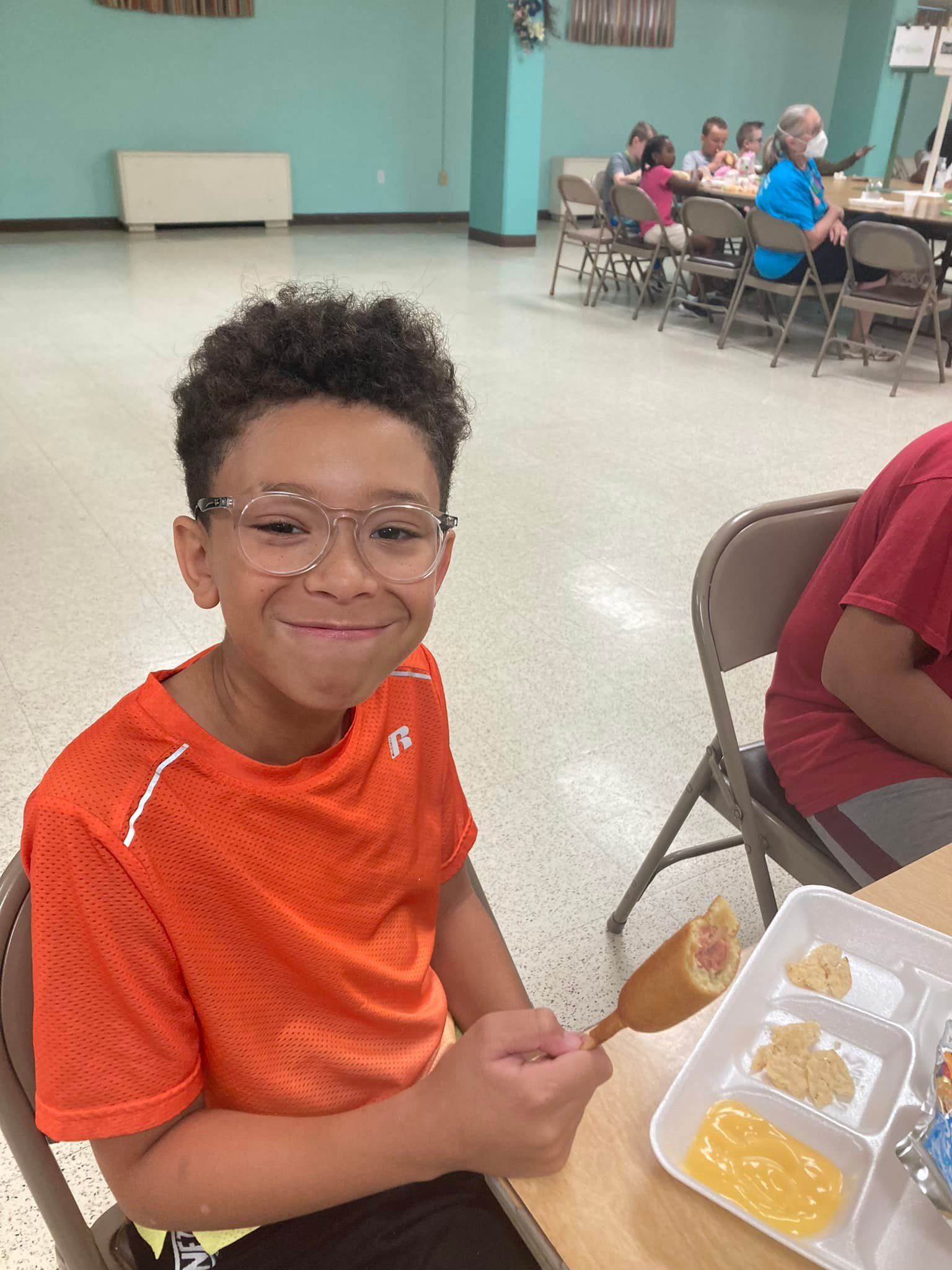 Smiling child in orange shirt seated at a table with trays of paint in a classroom, holding a brush