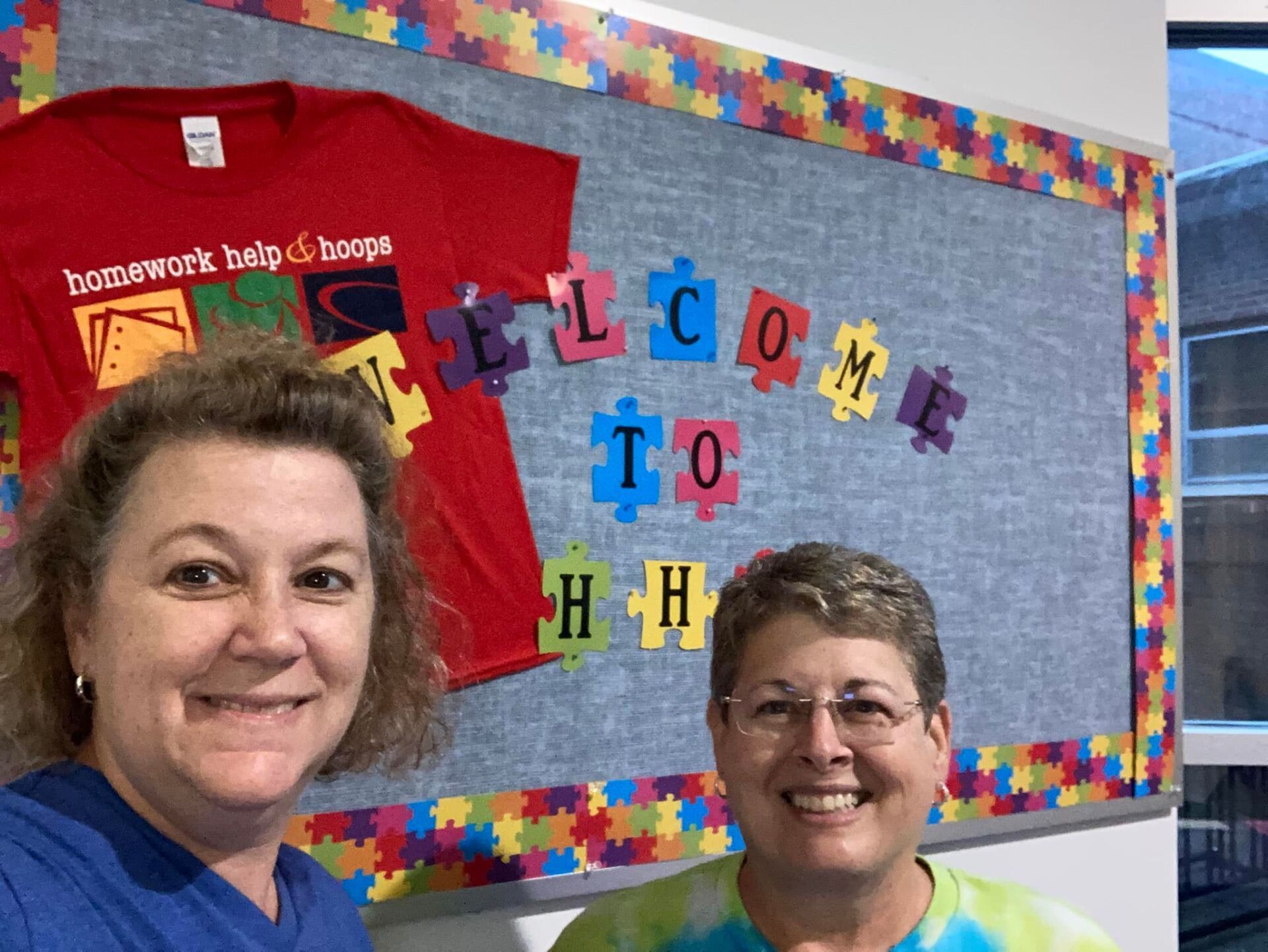 Two people smiling beside a colorful classroom bulletin board that says “Welcome to HH.”