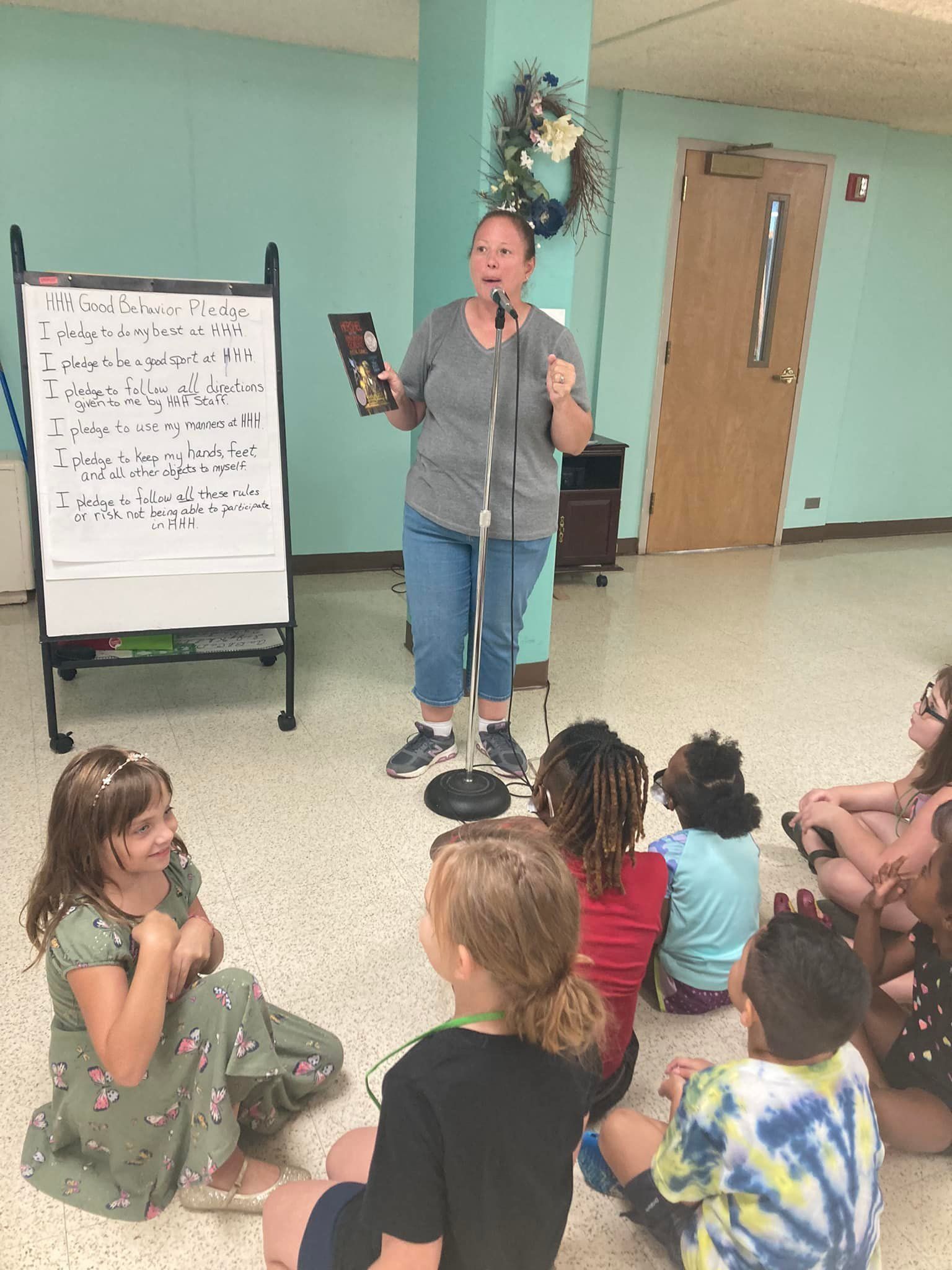 Presenter speaking to a seated group of children in a classroom, holding a book beside a flip chart.