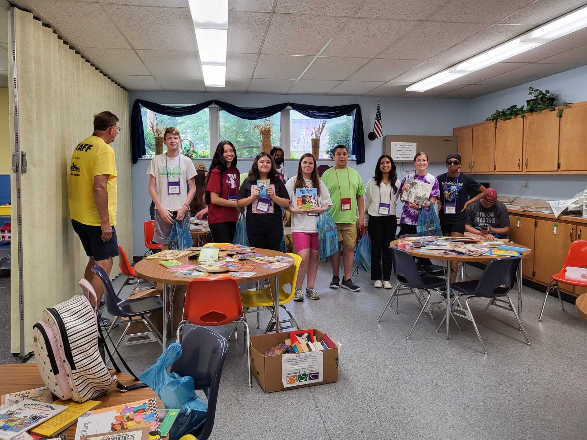 Group of people standing in a classroom around tables with snacks and crafts, preparing for an event.