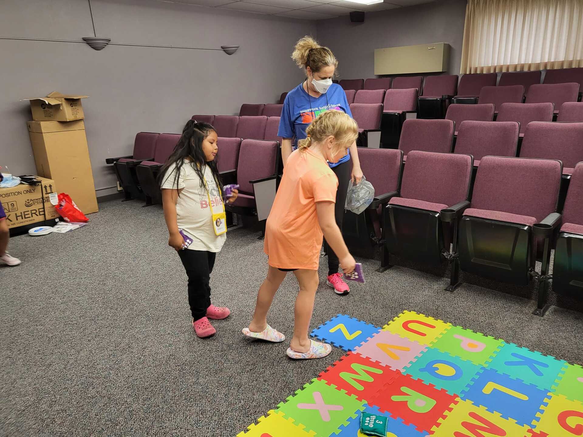 Children standing by colorful foam letter mat in a classroom or playroom, with tiered seating in the background