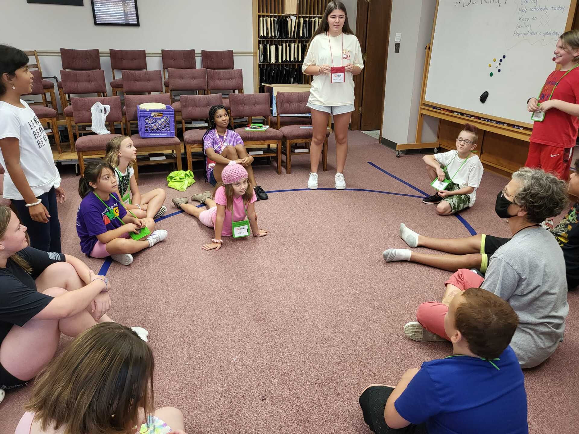 Children and adults sit in a circle on a carpeted room floor during a group activity.