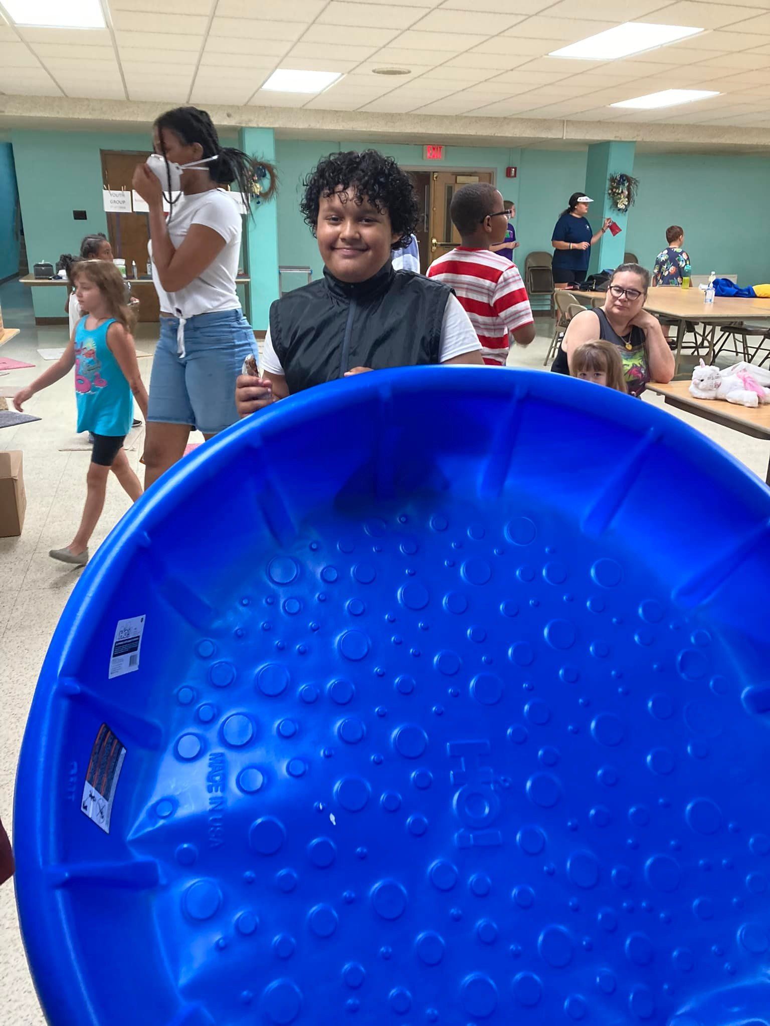 Person smiling and holding a large blue plastic basin in a community pool area