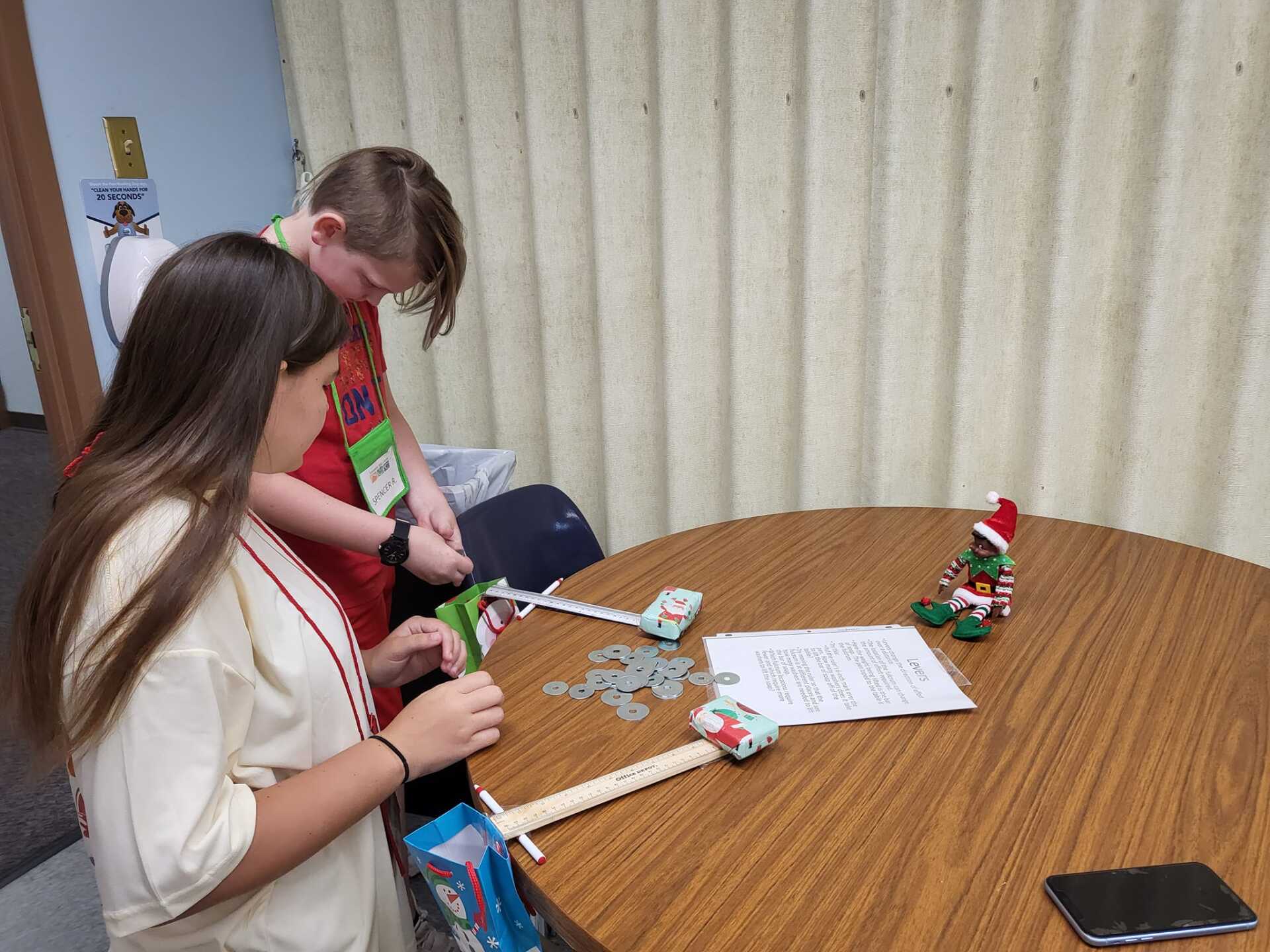 Two people craft at a round table with supplies, paper, and a small figurine; a phone lies nearby.
