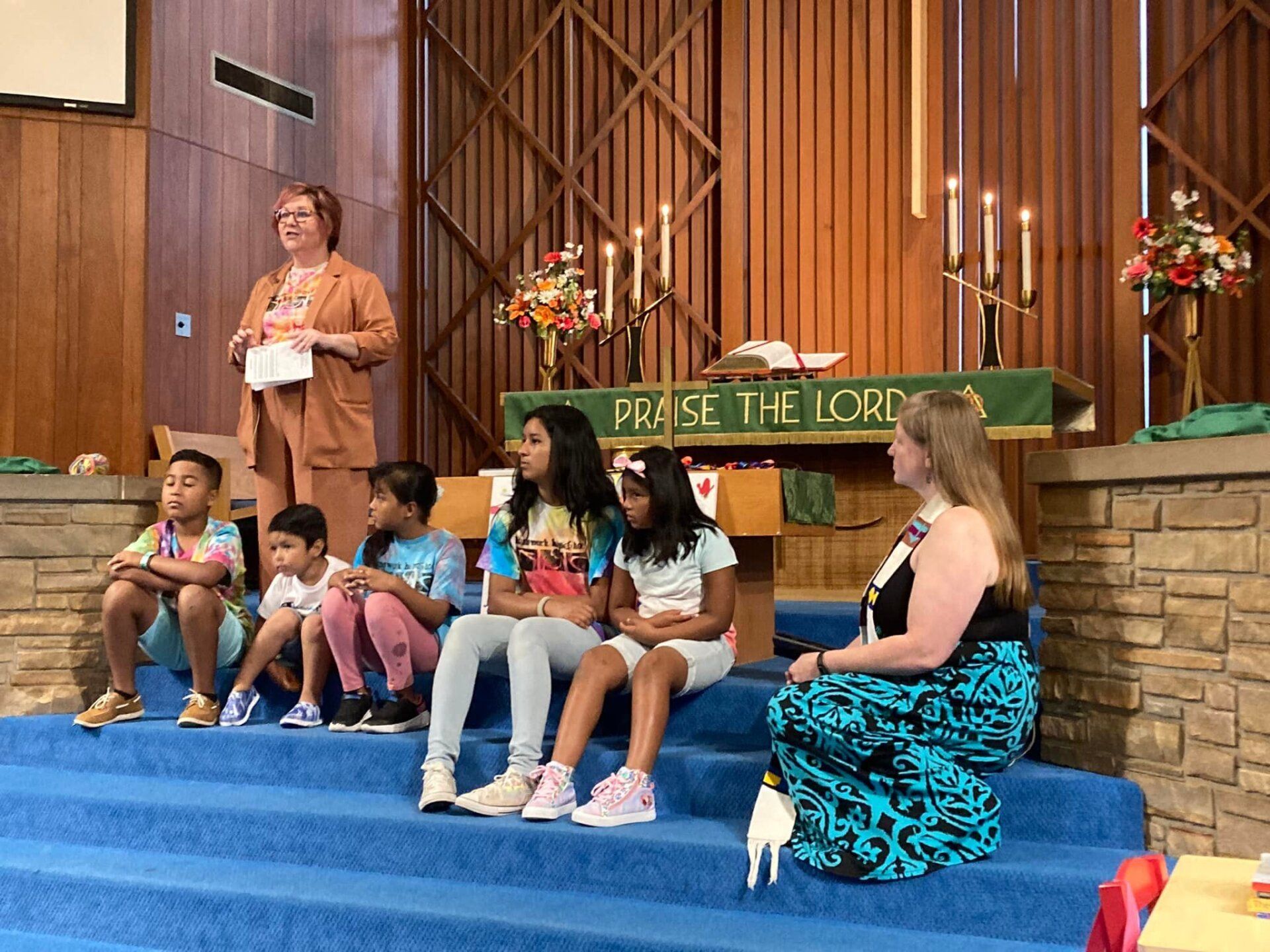 Children sit on a church stage while an adult speaks near a wooden cross and floral arrangements.