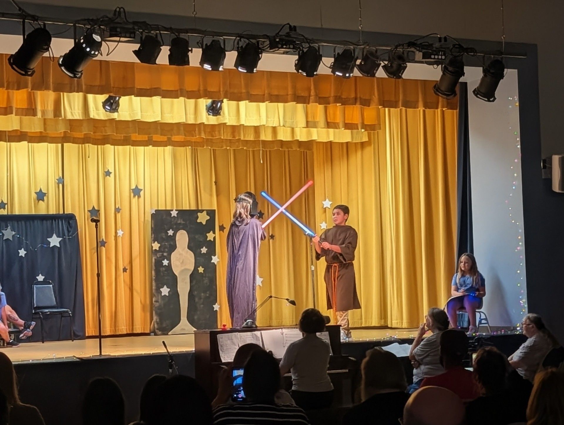 Children on a stage performing with glowing toy swords under yellow curtains, audience seated below