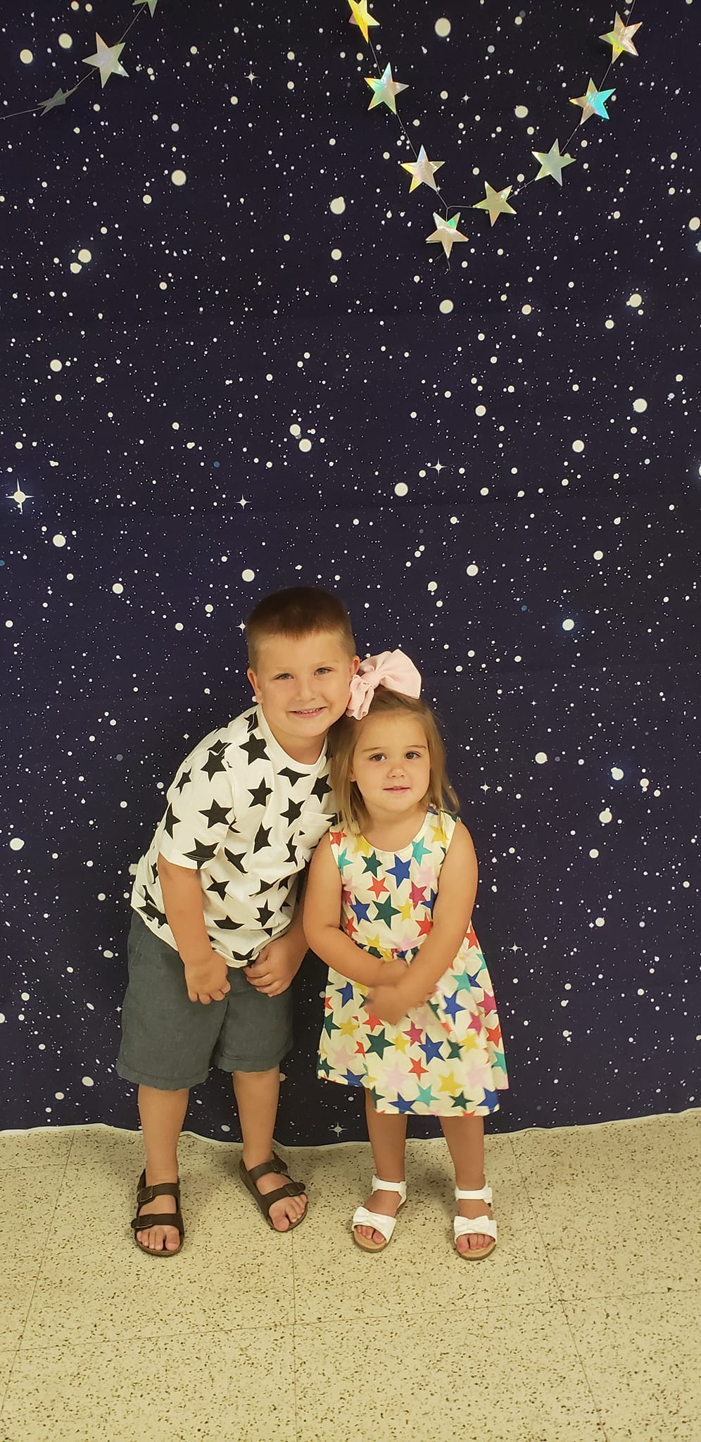 Two children standing together in front of a starry backdrop, smiling at the camera.