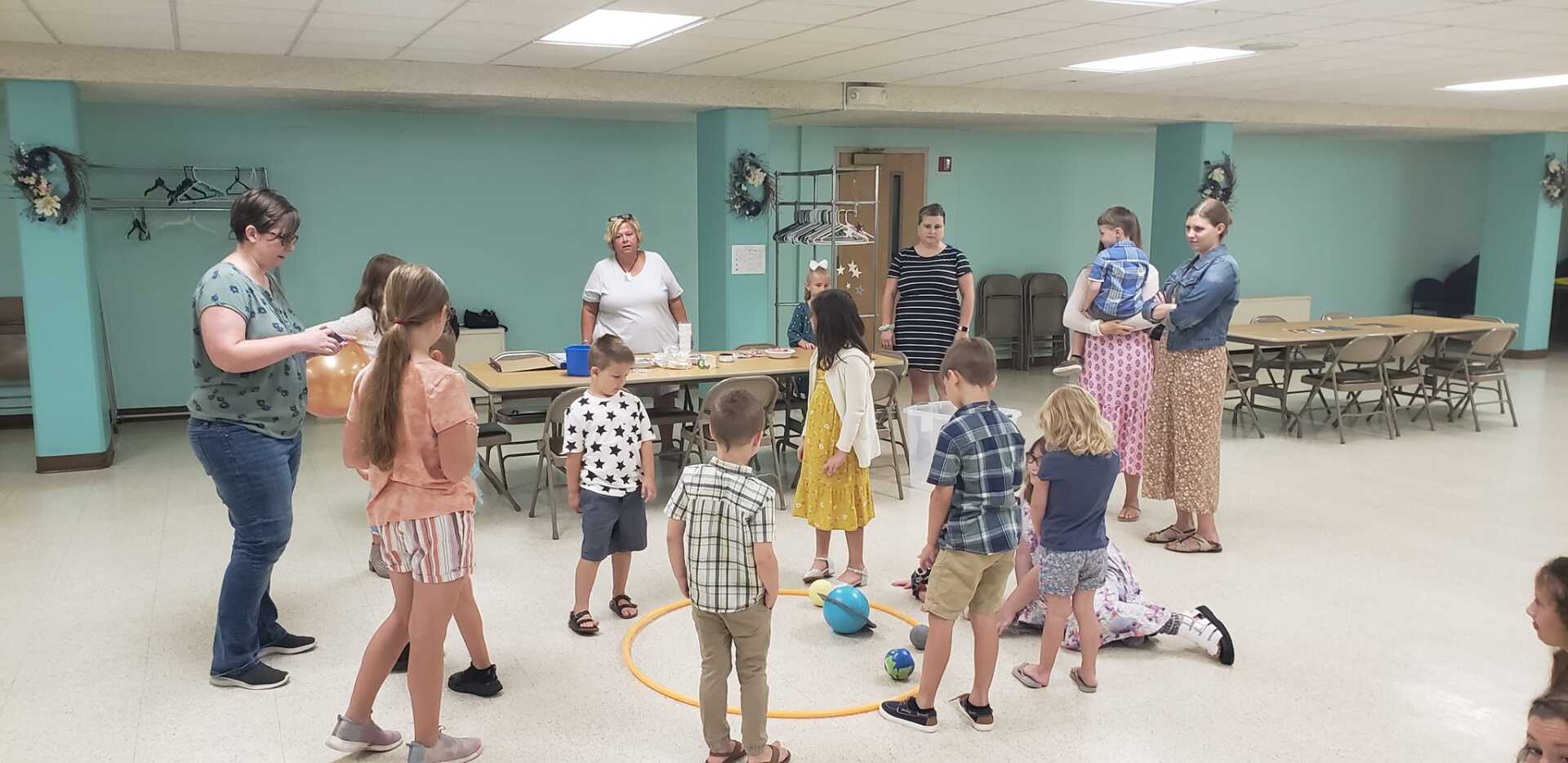 People gathered in a bright room, with children standing in a circle around a table and adults nearby.