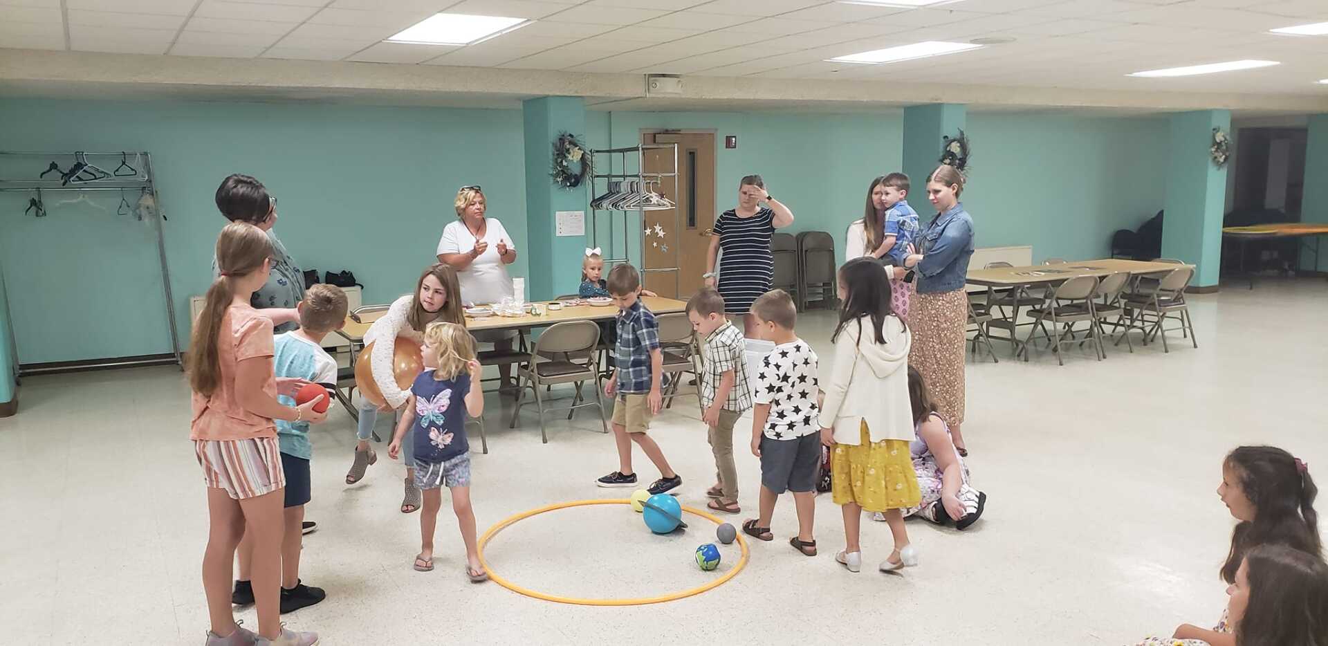 Children gathered in a gym around a yellow hoop, with adults supervising in the background.