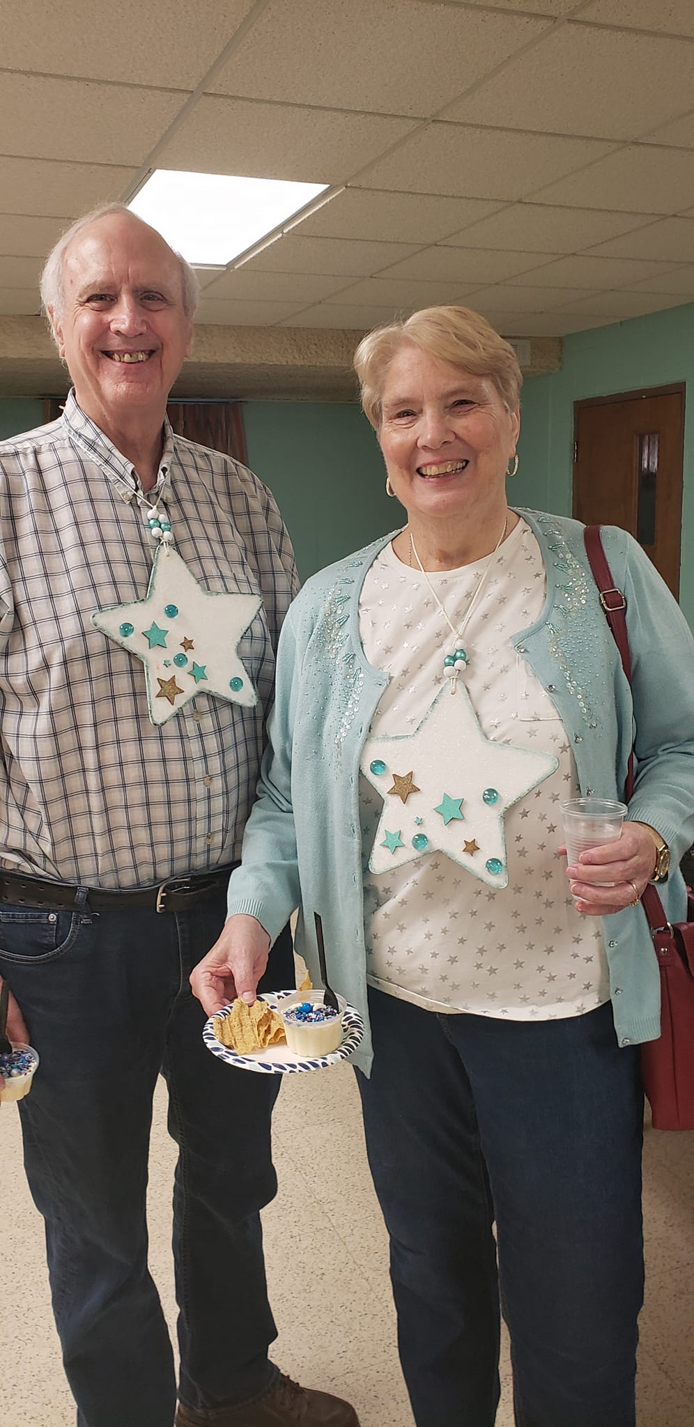 Two people in a hallway, smiling and holding plates at an indoor gathering.
