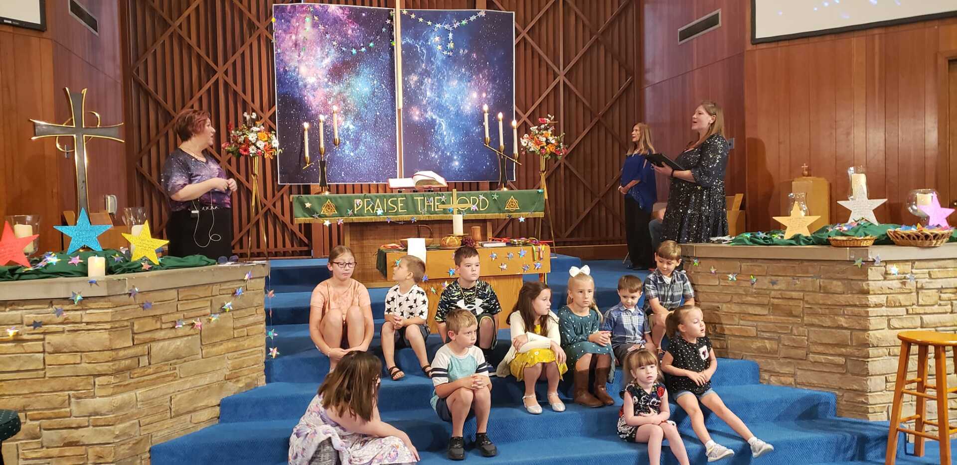 Children seated on a church stage with musicians performing beside a starry backdrop