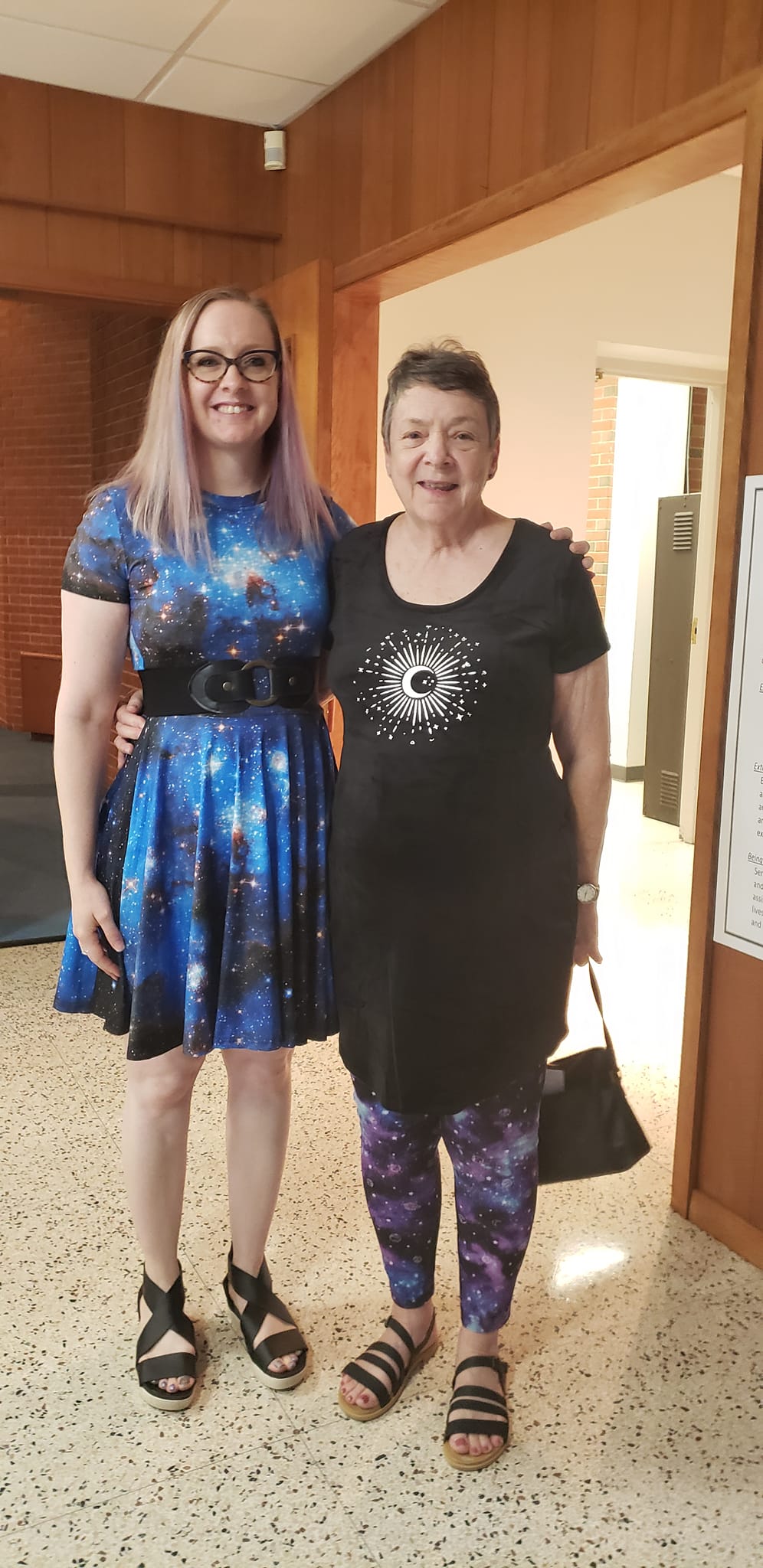 Two women standing together in a hallway, one in a blue dress and one in a black dress with purple leggings.