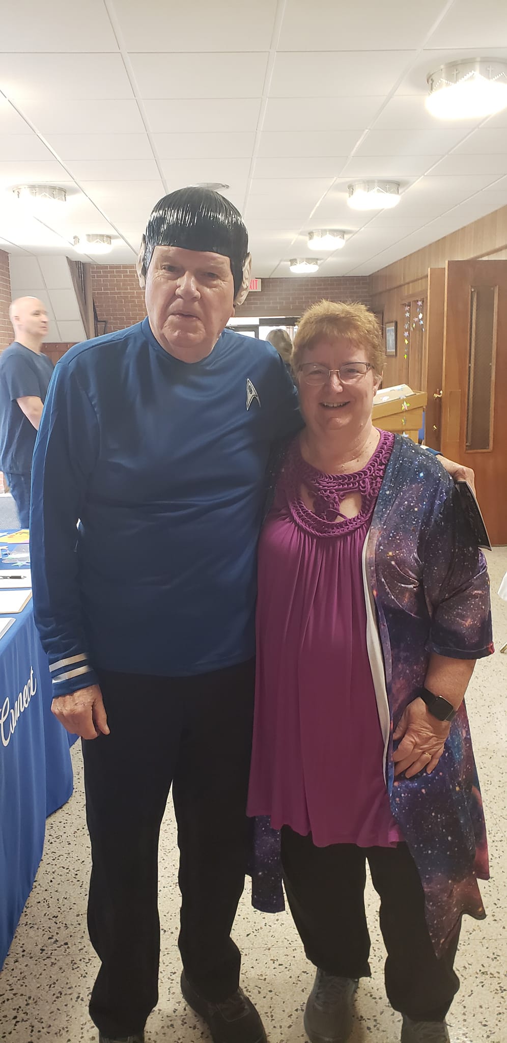 Two people posing indoors, smiling, with a blue table and event booths in the background.