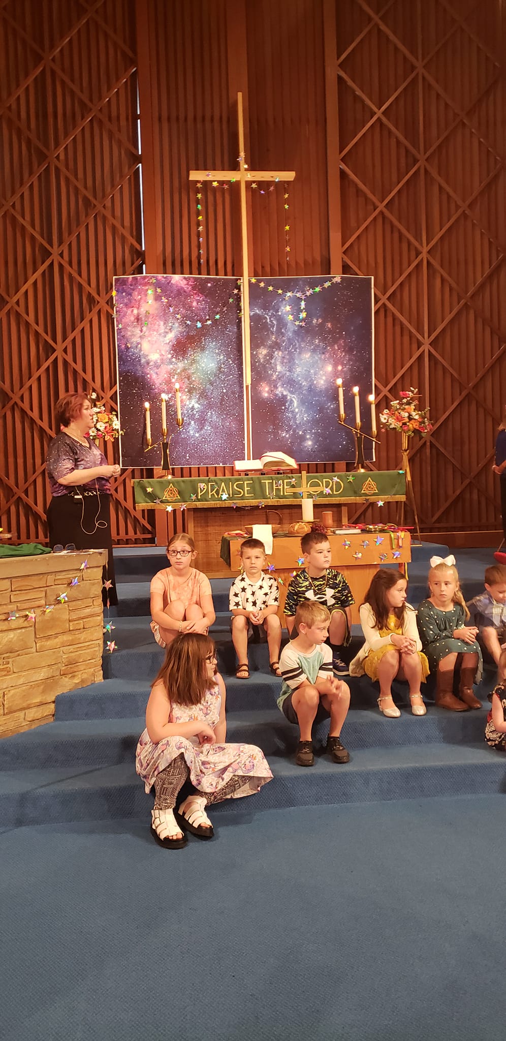 People seated on blue carpet before a church altar with a cross and candles, and a starry backdrop.