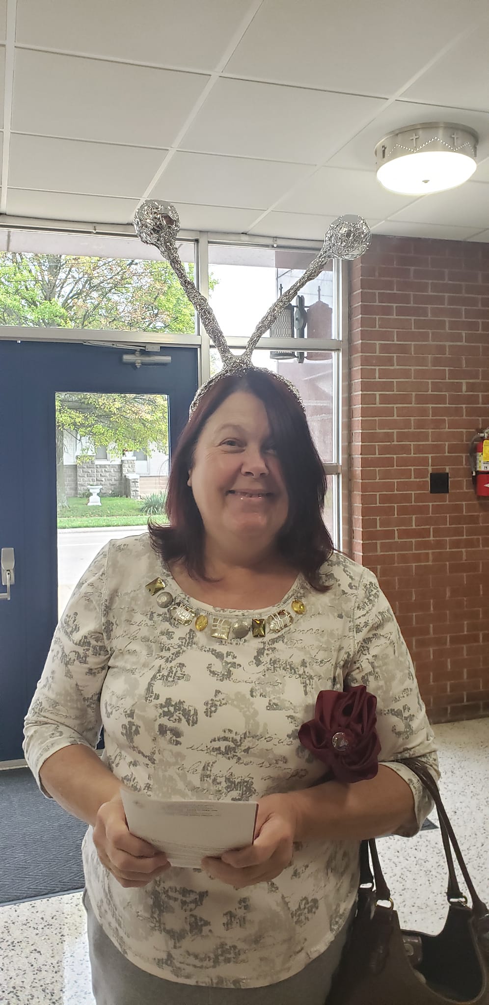 Woman in a white floral top holds papers in a lobby with brick walls and a blue door.