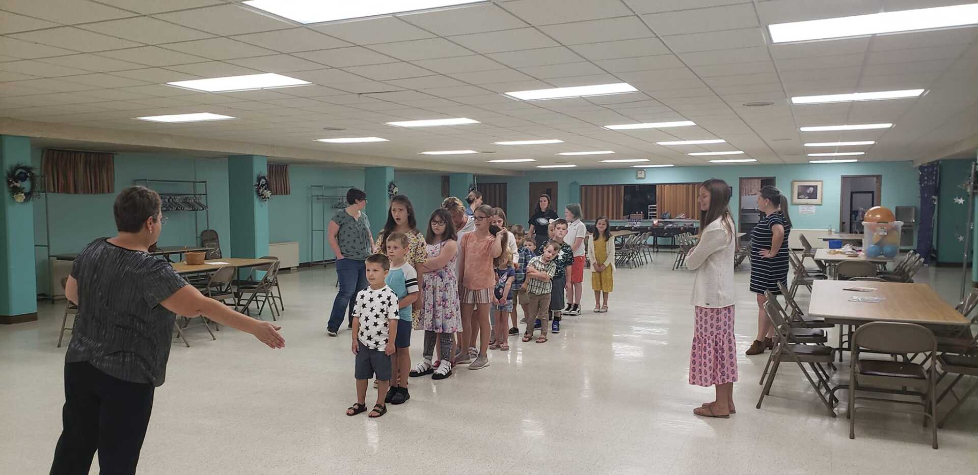 People lined up in a bright hall during a group activity, with a leader gesturing at left and tables along the right.