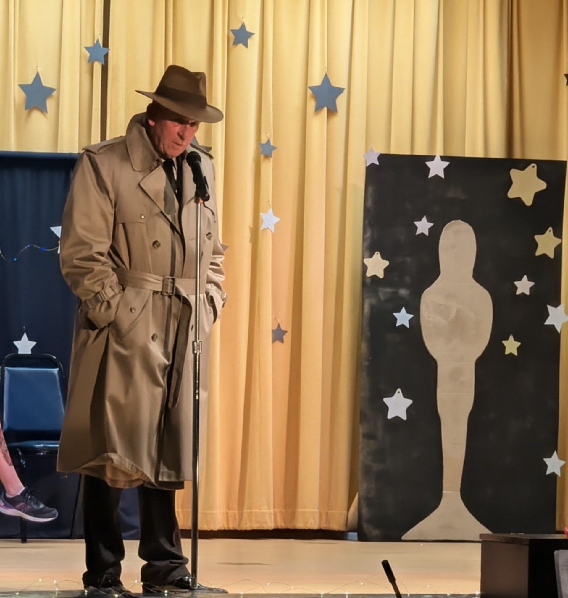 Man in trench coat and hat stands on stage beside a silhouette target and starry backdrop