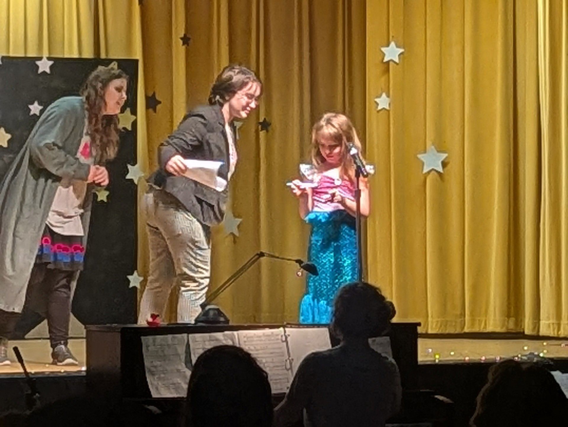 Three people on stage with microphones, performing in front of gold curtains and star decorations.