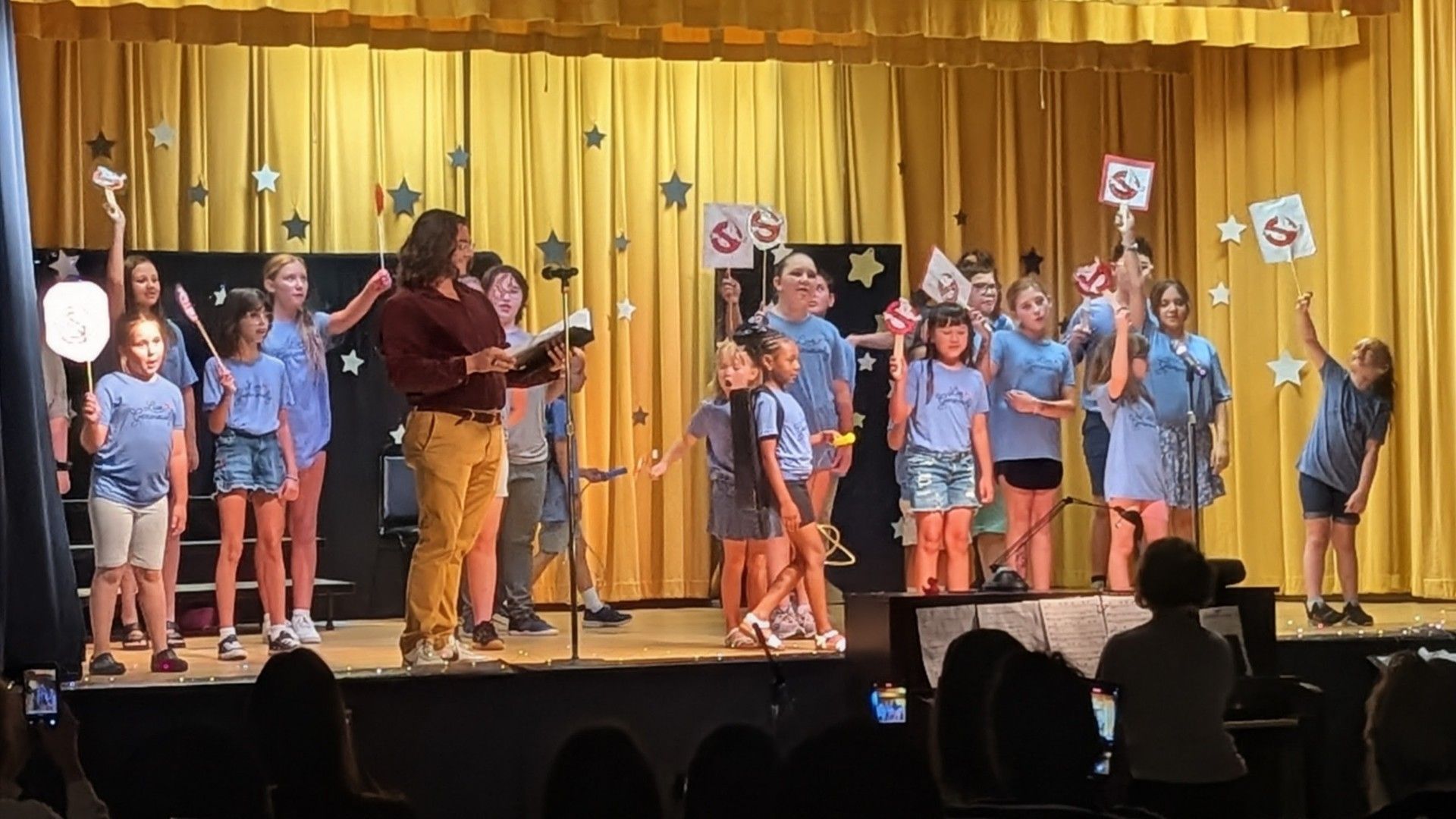 School choir students perform onstage with microphones in front of a gold curtain.